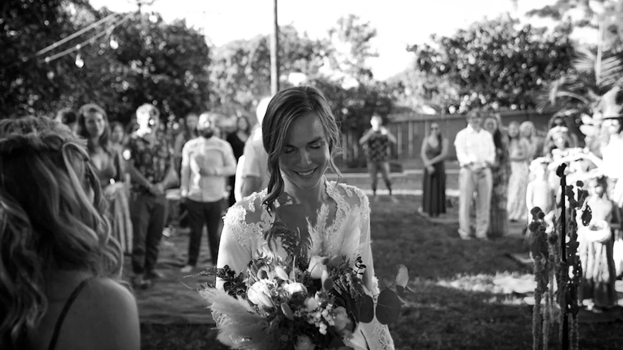 A woman in a white lace dress holding a bouquet of flowers at an outdoor event, with guests in the background.