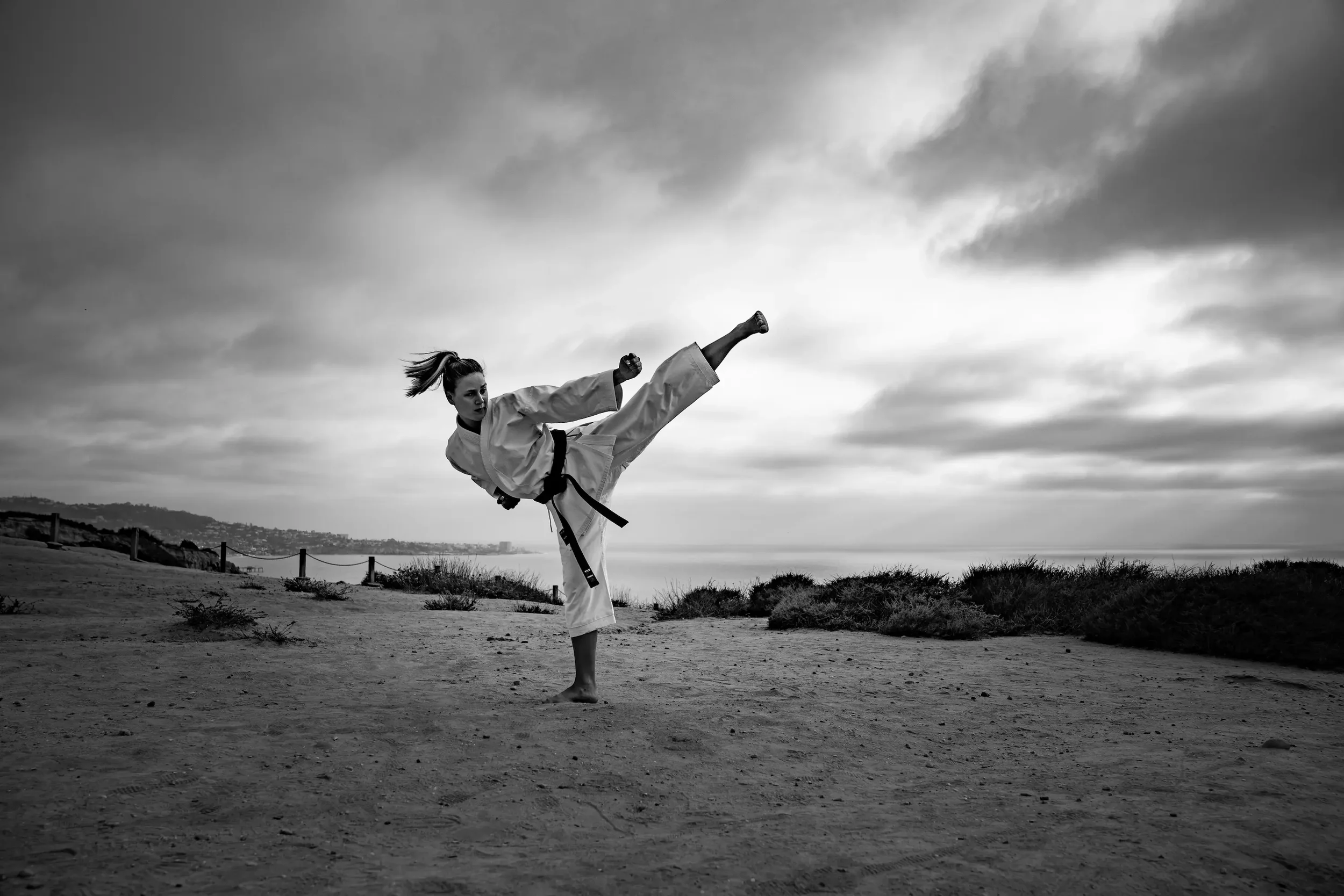 A woman practicing martial arts outdoors on a sandy area under a cloudy sky, wearing a traditional martial arts uniform and a black belt, performing a high kick.