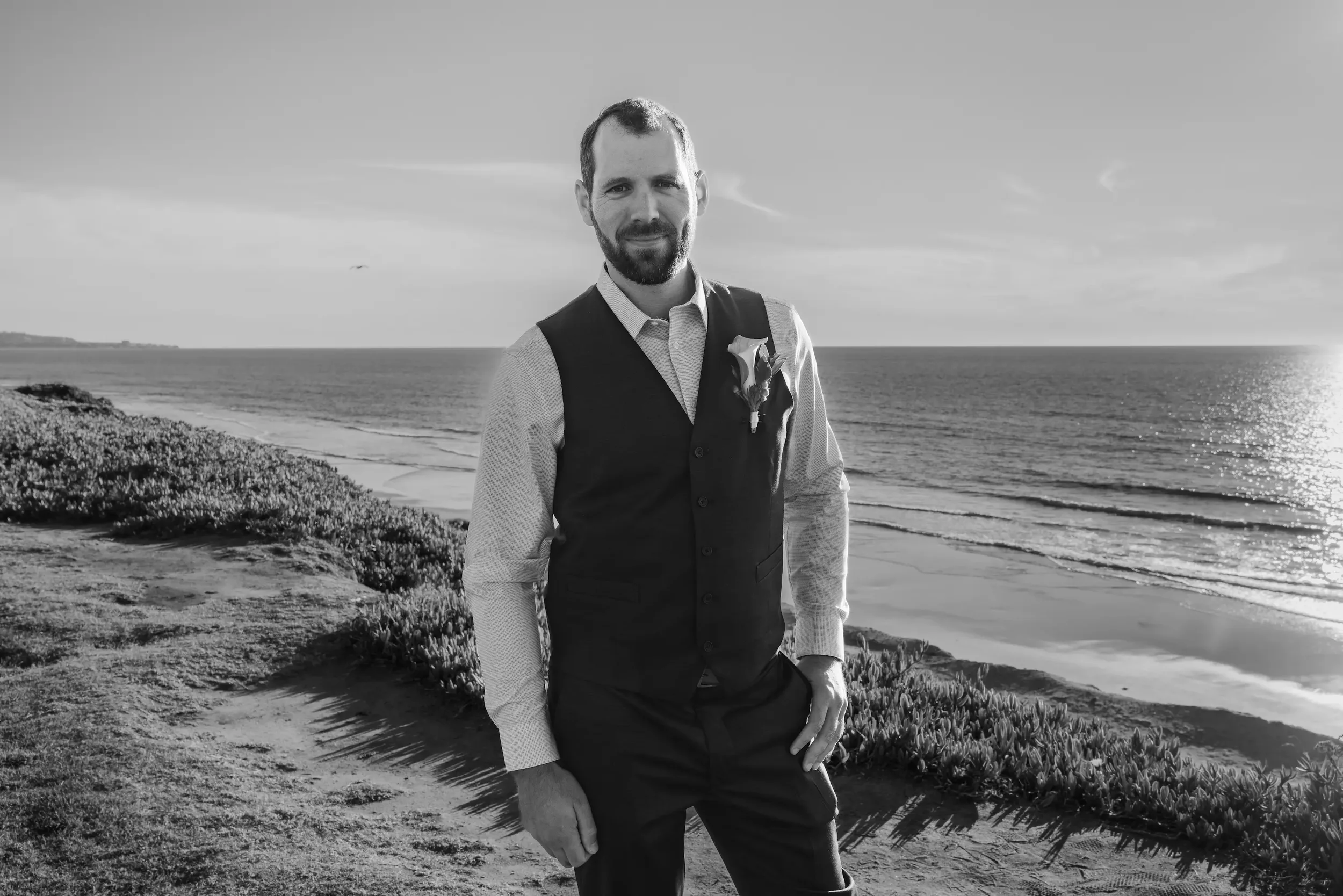 Black and white photo of a man with a beard and short hair standing on a beach, wearing a dress shirt, vest, and pocket square, with ocean and sky in the background.