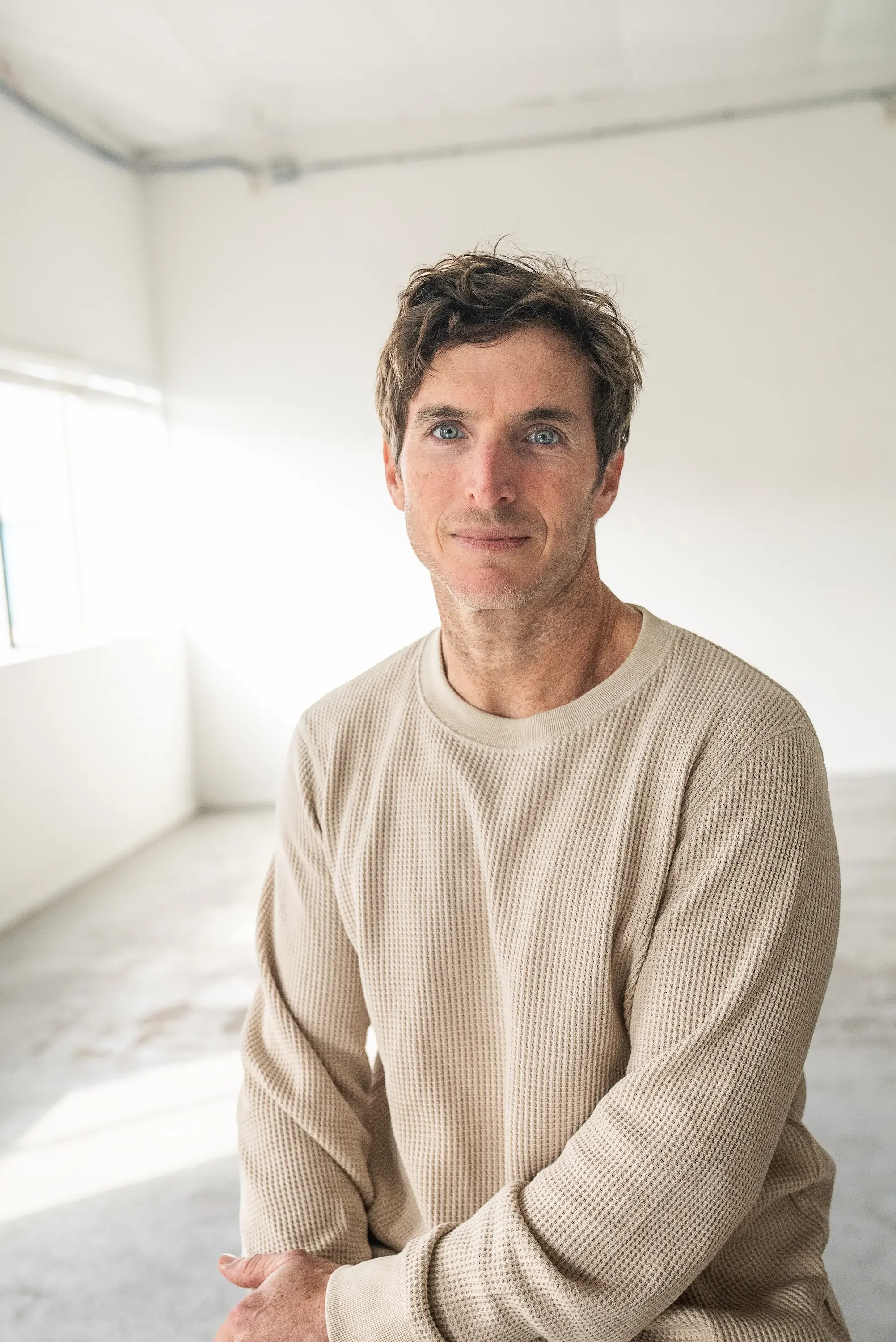A young man with curly brown hair and blue eyes, wearing a beige sweater, sitting in a bright, minimalist room with white walls and large windows.
