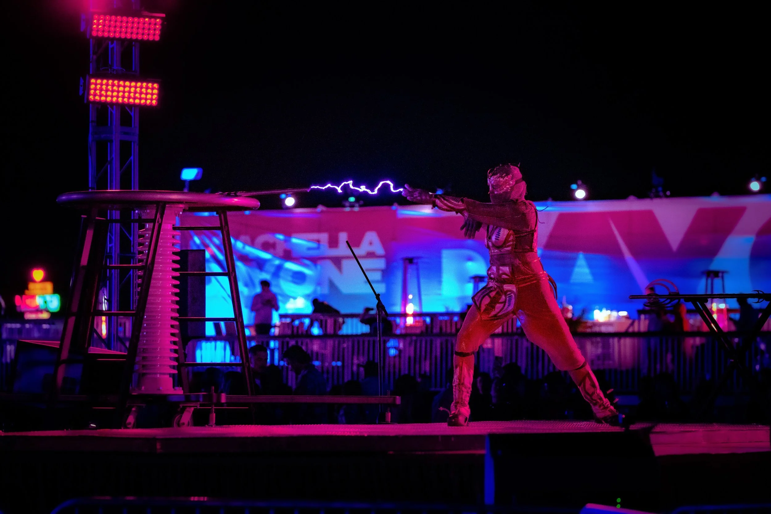 A person dressed in a costume creates a spark with a Tesla coil at night, illuminated by colorful lights.