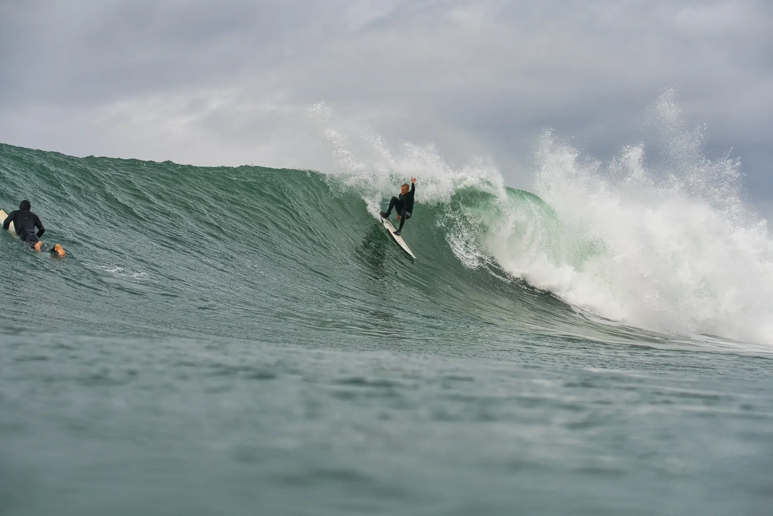 A man surfing on a wave in the ocean while another person waits in the water nearby.