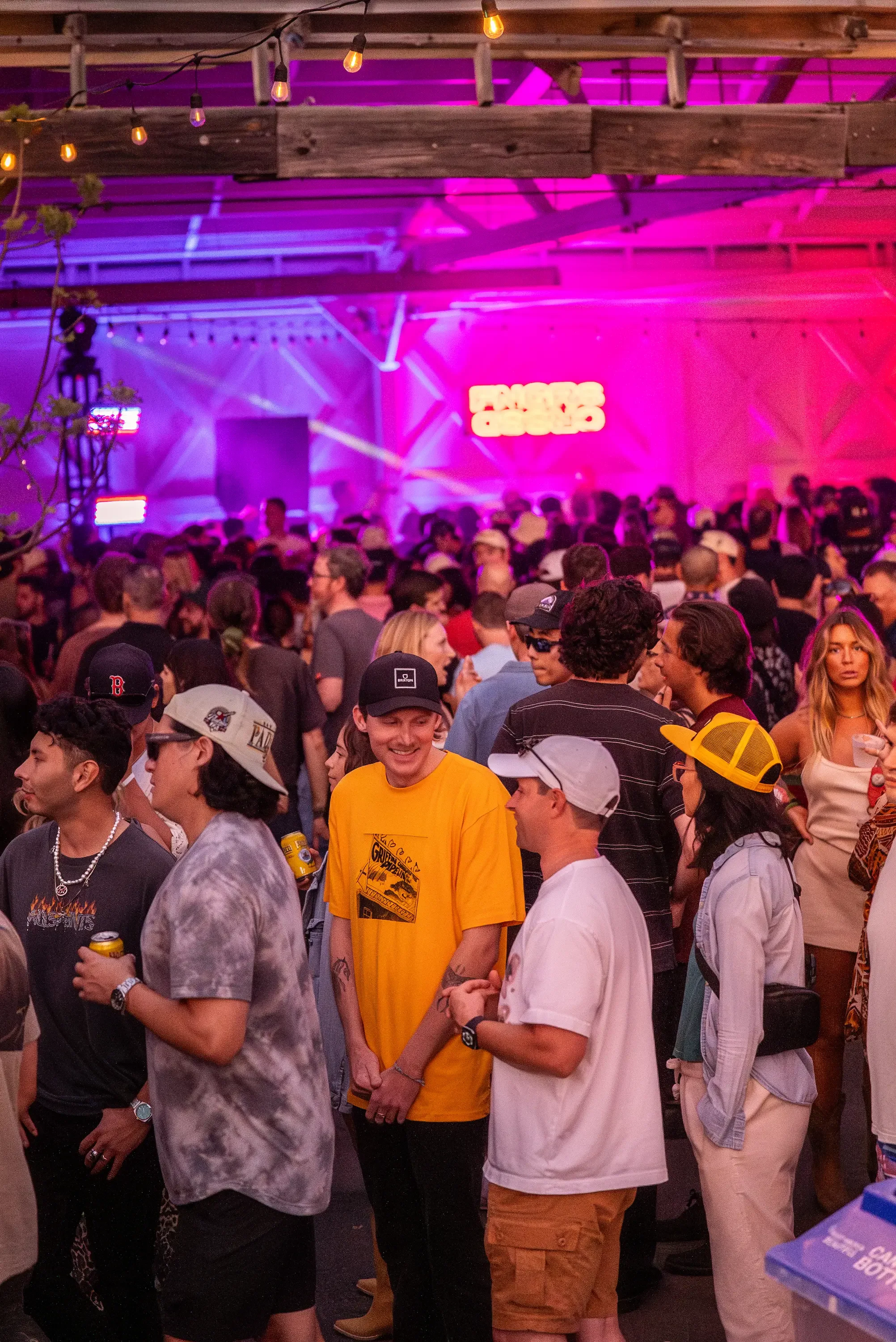 A crowded indoor event space with a purple and pink lighting, filled with people standing and socializing. There are string lights hanging from the ceiling, and a neon sign in the background. People are dressed casually, some with hats and sunglasses.