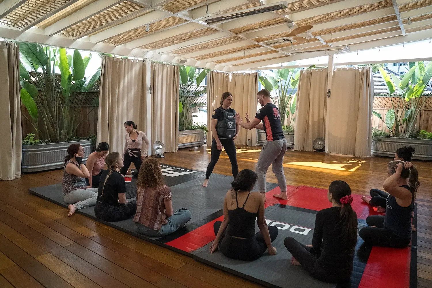 A group of women practicing martial arts or self-defense in a room with wooden floors and curtains, watching two instructors demonstrating techniques.