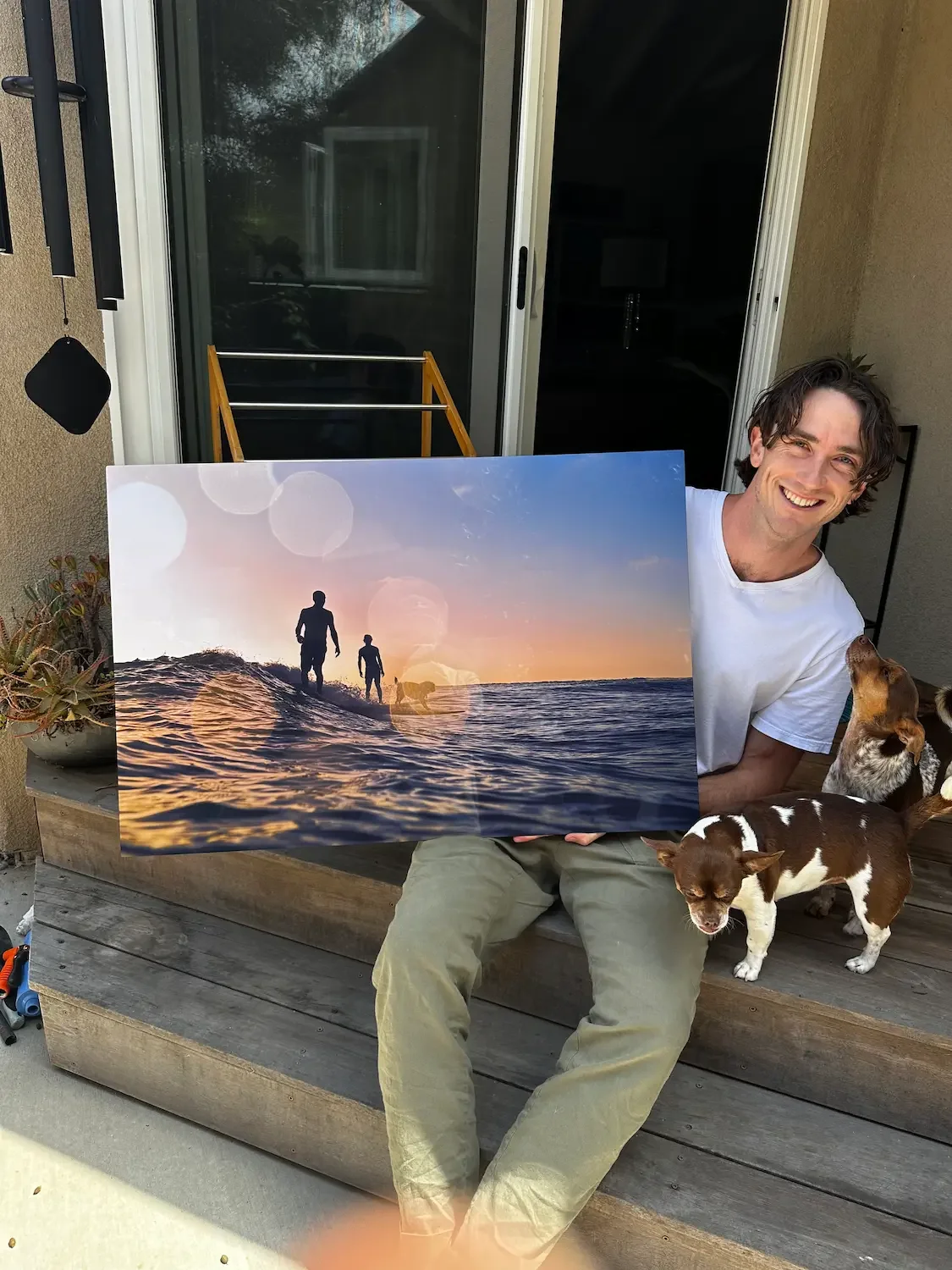 Jack Lajoie holding a large printed canvas of a surf photo, with two dogs beside him on a porch