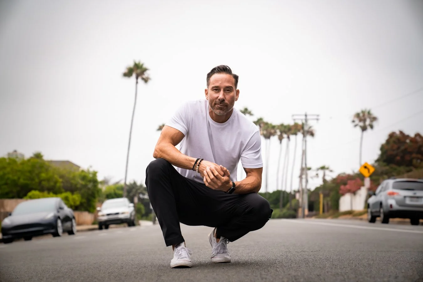 A man crouching on a street with palm trees and parked cars in the background.