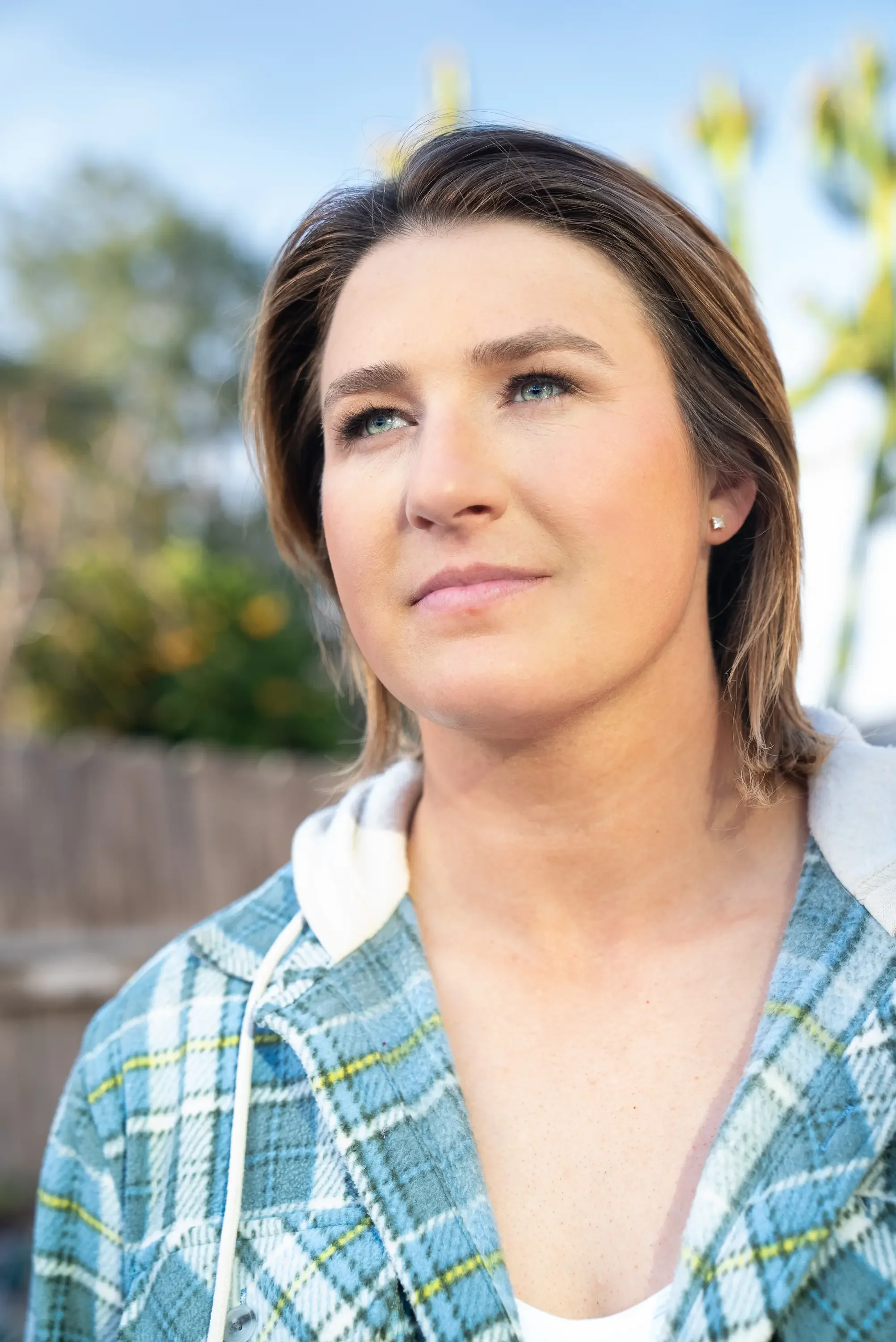A woman with short brown hair and blue eyes, wearing a blue and green plaid hoodie with a white inner shirt, outdoors with blurred trees and sky in the background.