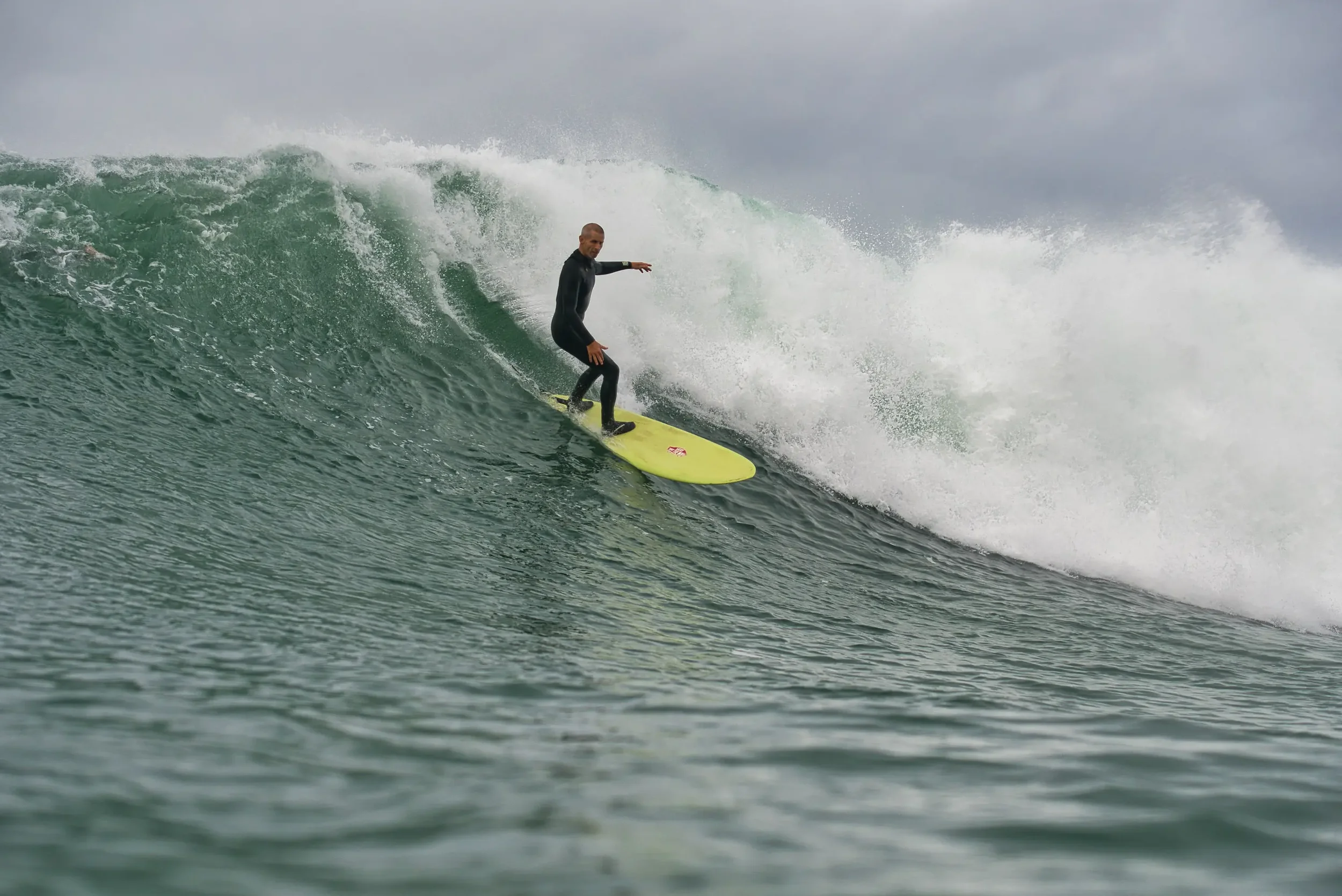 A person in a black wetsuit surfing on a large wave with a cloudy sky in the background.
