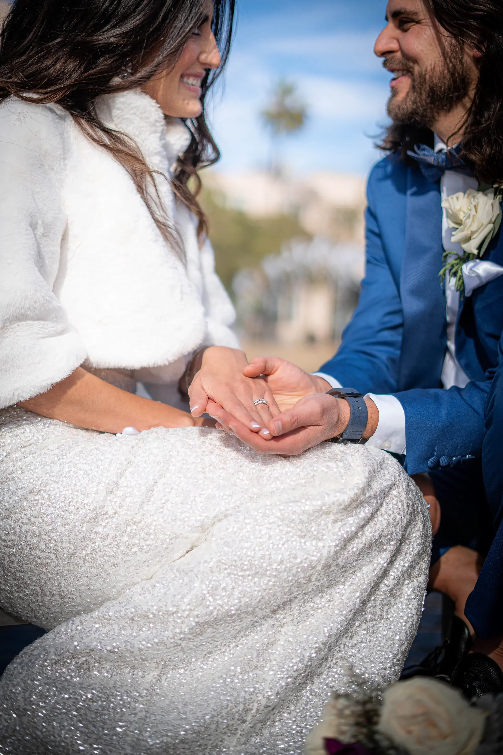 A close-up of a couple at their wedding, holding hands and smiling at each other. The bride is wearing a sparkly white wedding gown and a white fur shawl, and the groom is dressed in a blue suit with a white shirt, navy bow tie, and boutonnière.