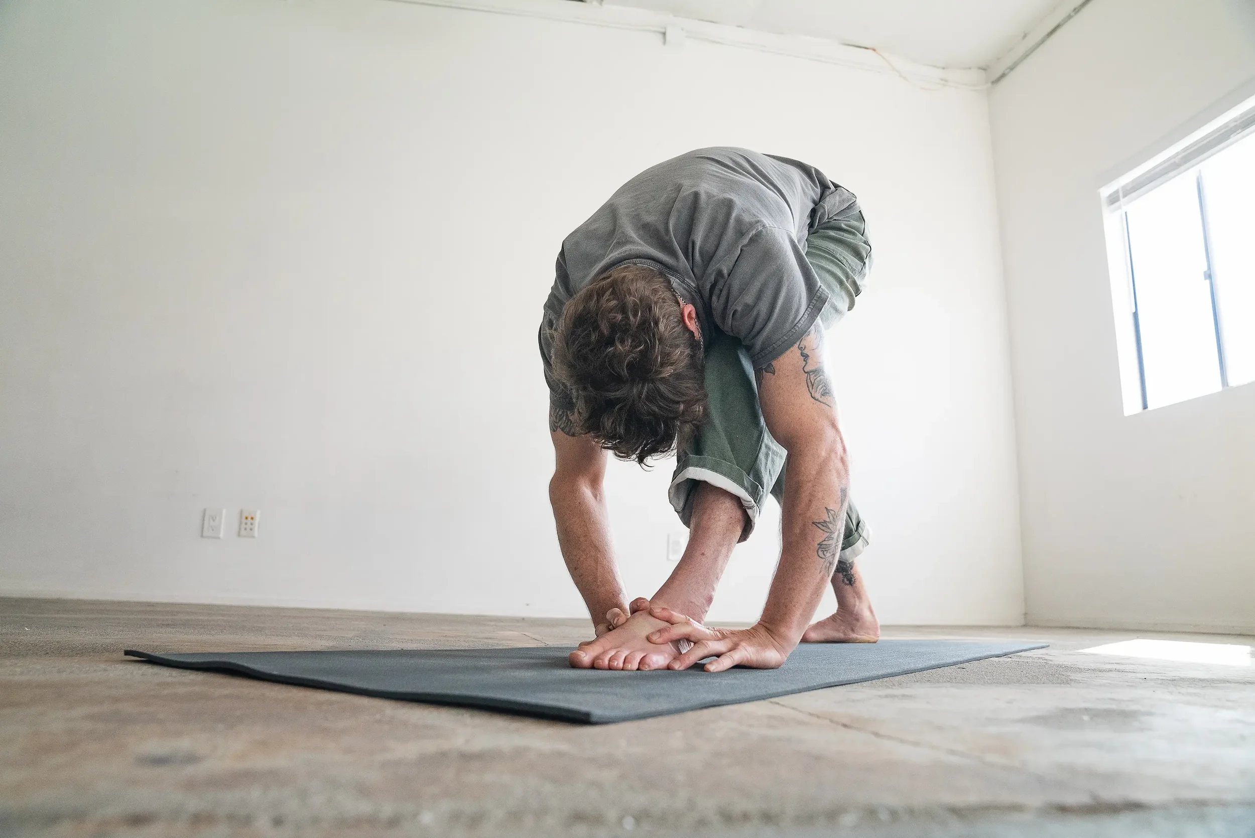 A person with tattoos practicing yoga indoors on a yoga mat, balancing on one hand and one foot in a downward dog pose, with a plain white wall and window in the background.