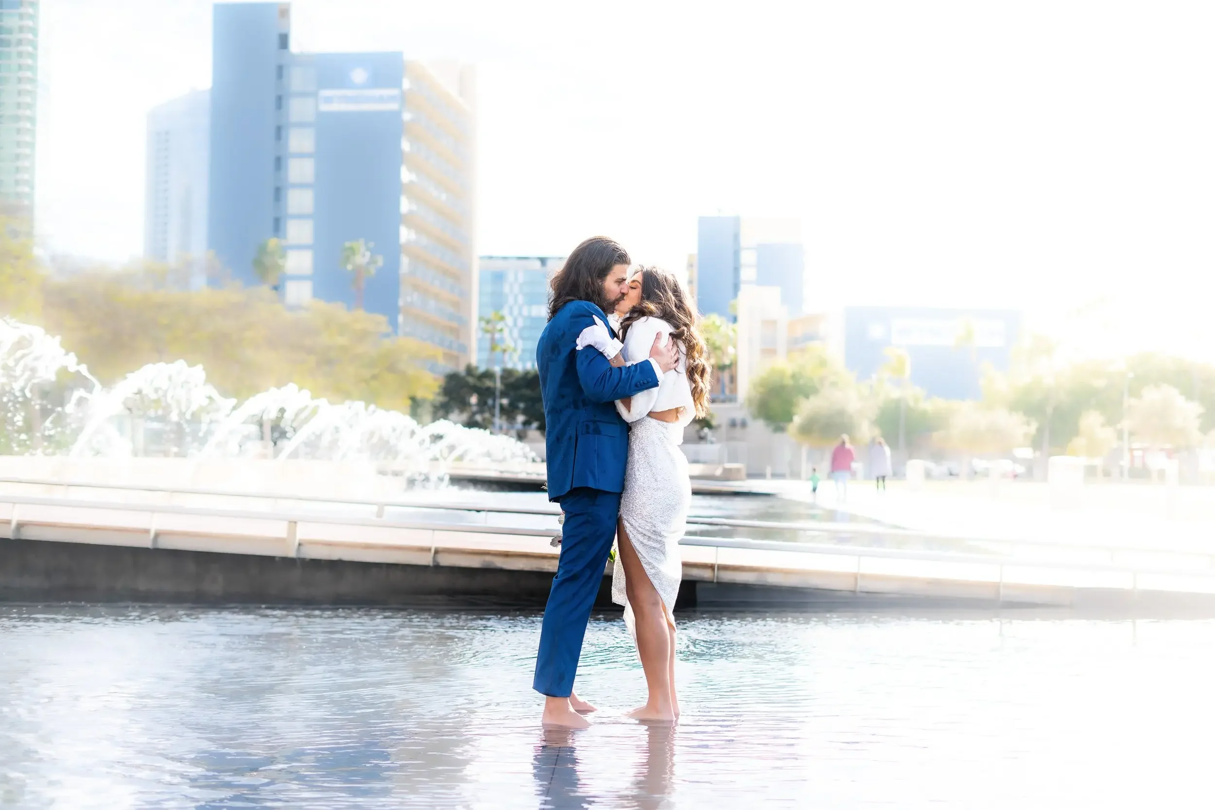 A couple dressed in formal attire sharing a kiss while standing in a shallow fountain in an urban park with tall buildings in the background.