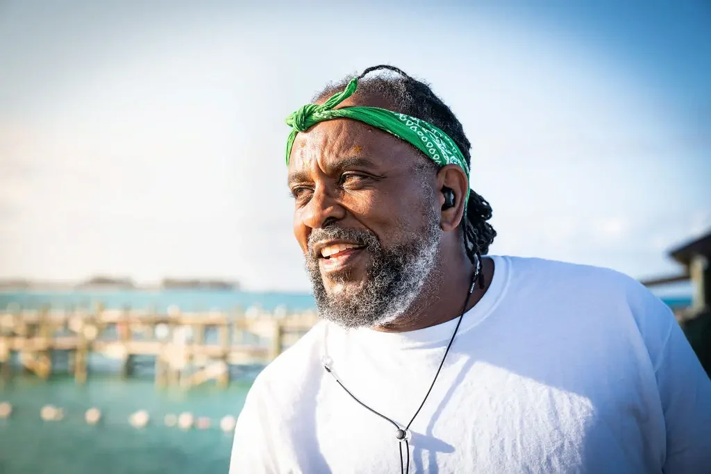 A middle-aged man with a beard and dreadlocks wearing a green bandana, a white t-shirt, and earbuds, standing outdoors near a body of water with a dock in the background on a sunny day.