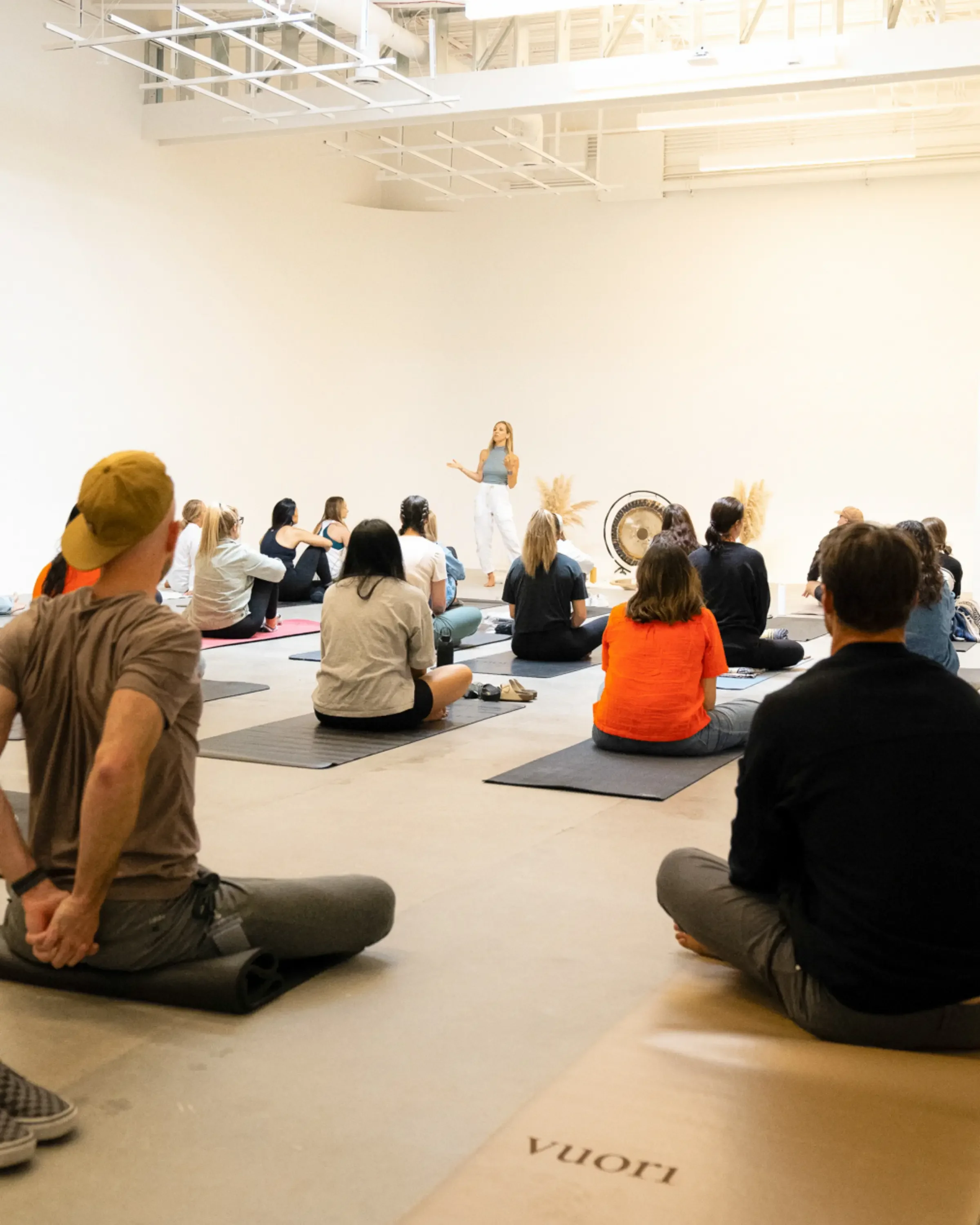 A group of people sitting on yoga mats in a spacious, well-lit room, listening to a woman standing and speaking in front of them. Some of the attendees have notebooks and water bottles. The room has a minimalist design with a high ceiling, and there is a large gong and decorative pampas grass in the background.