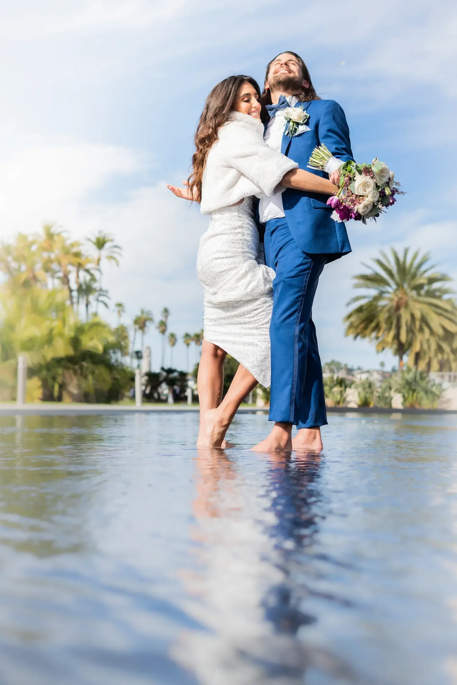 A bride and groom are happily standing in shallow water at a beach or lakeside, embracing each other. The bride is wearing a white dress and a white shawl, and the groom is dressed in a bright blue suit with a white shirt, bow tie, and a white flower boutonniere. The groom is holding a bouquet of flowers, and both are smiling with their eyes closed, enjoying a joyful moment with palm trees and a partly cloudy sky in the background.