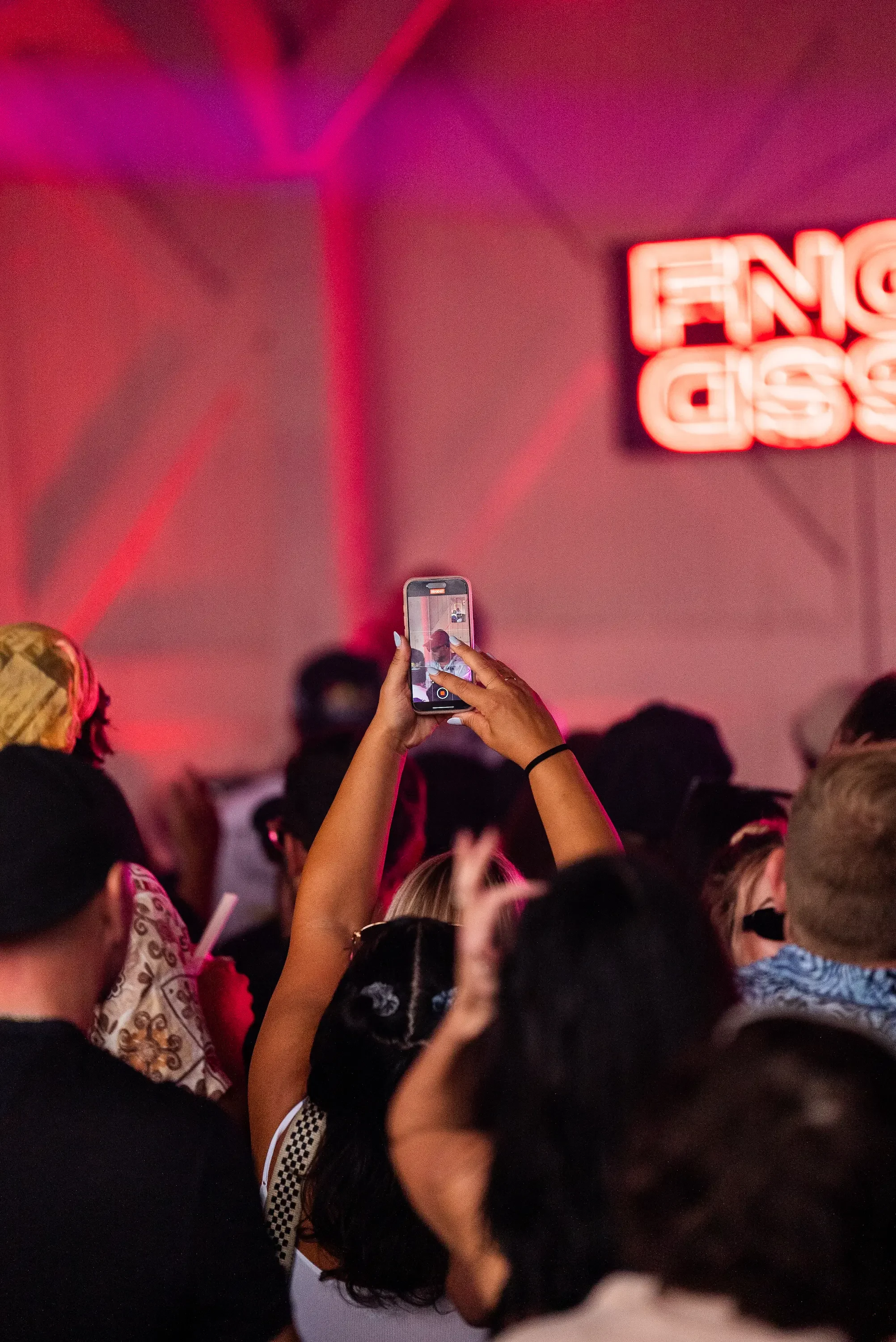 A person is holding a smartphone taking a photo or video of a crowd of people at an event with pink and purple lighting and a neon sign reading "ENGLISH" in the background.