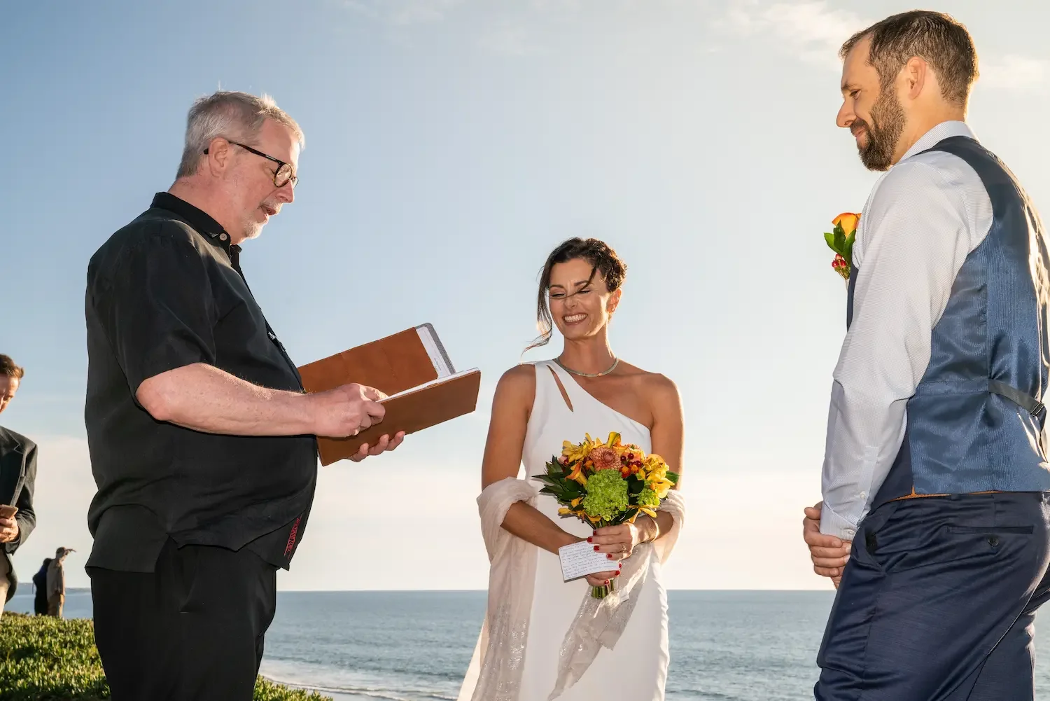 Officiant reading from a book as a bride and groom exchange vows with the ocean behind them