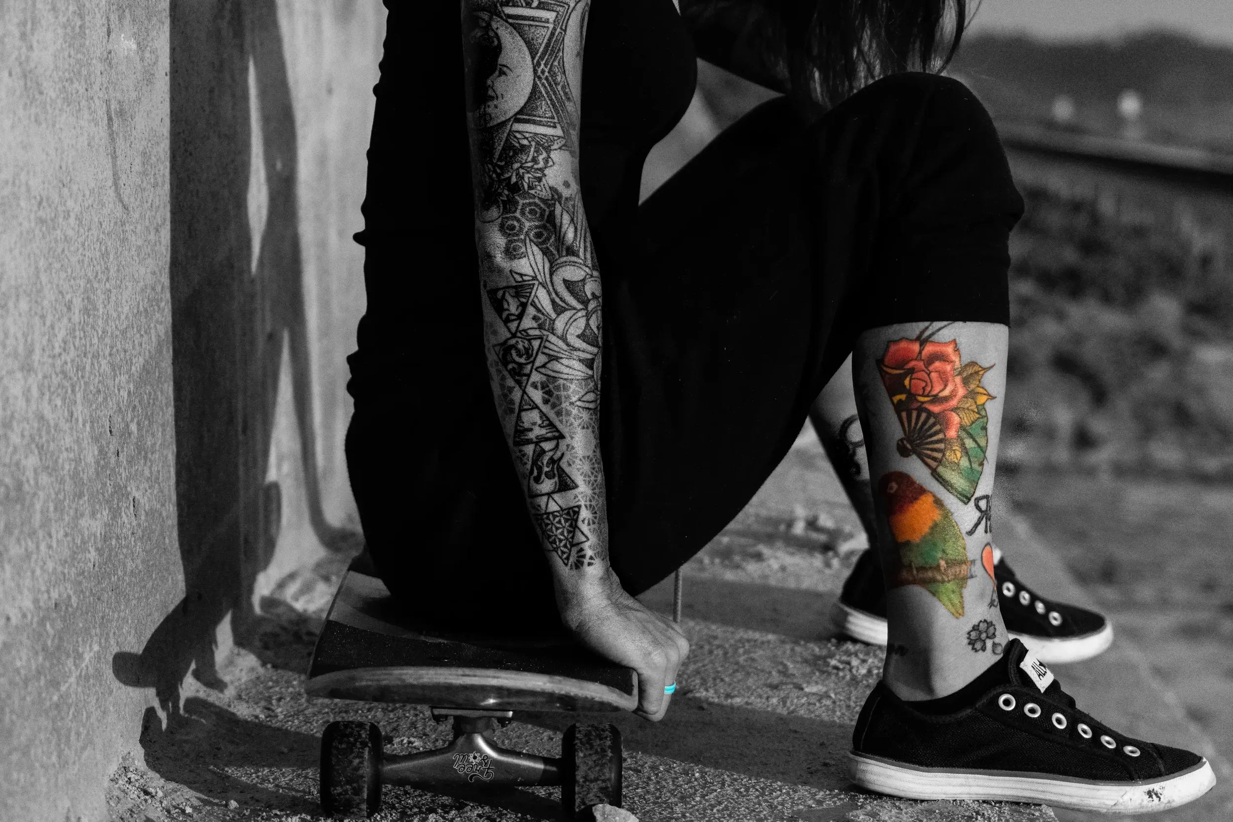 A person with tattoos on their arms sitting on a skateboard against a wall. The person is wearing black shoes and black clothing. The tattoos include colorful designs of a rose and other patterns.