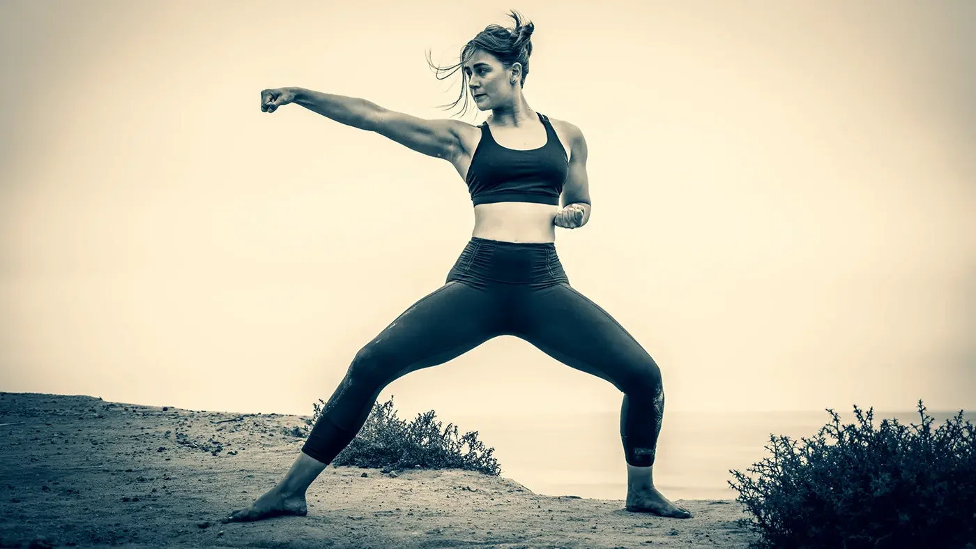 A woman in activewear practicing a martial arts pose outdoors on a dirt path with bushes, in black and white