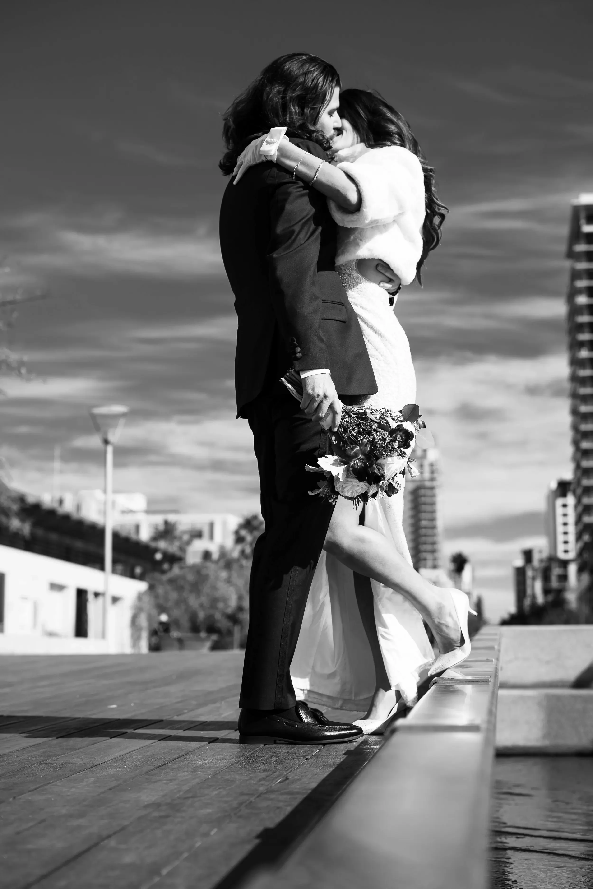A black and white photo of a couple embracing on a rooftop, with an urban cityscape in the background. The woman is dressed in a wedding gown, and the man is in a suit. She is holding a bouquet of flowers, and they are standing close, sharing an intimate moment.