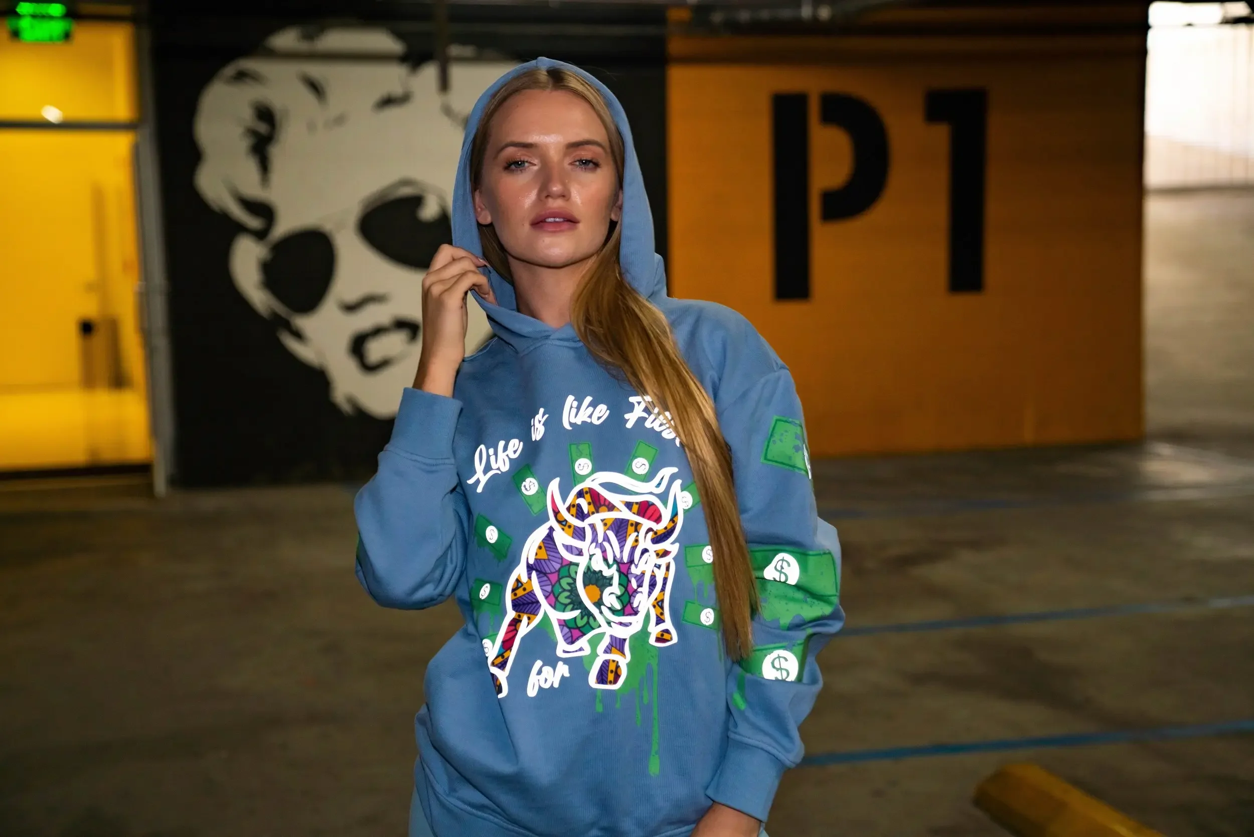 A young woman with long red hair wearing a blue hoodie with colorful graphic design stands in an indoor parking garage with yellow and black safety barriers and a black and white portrait of a person in the background.