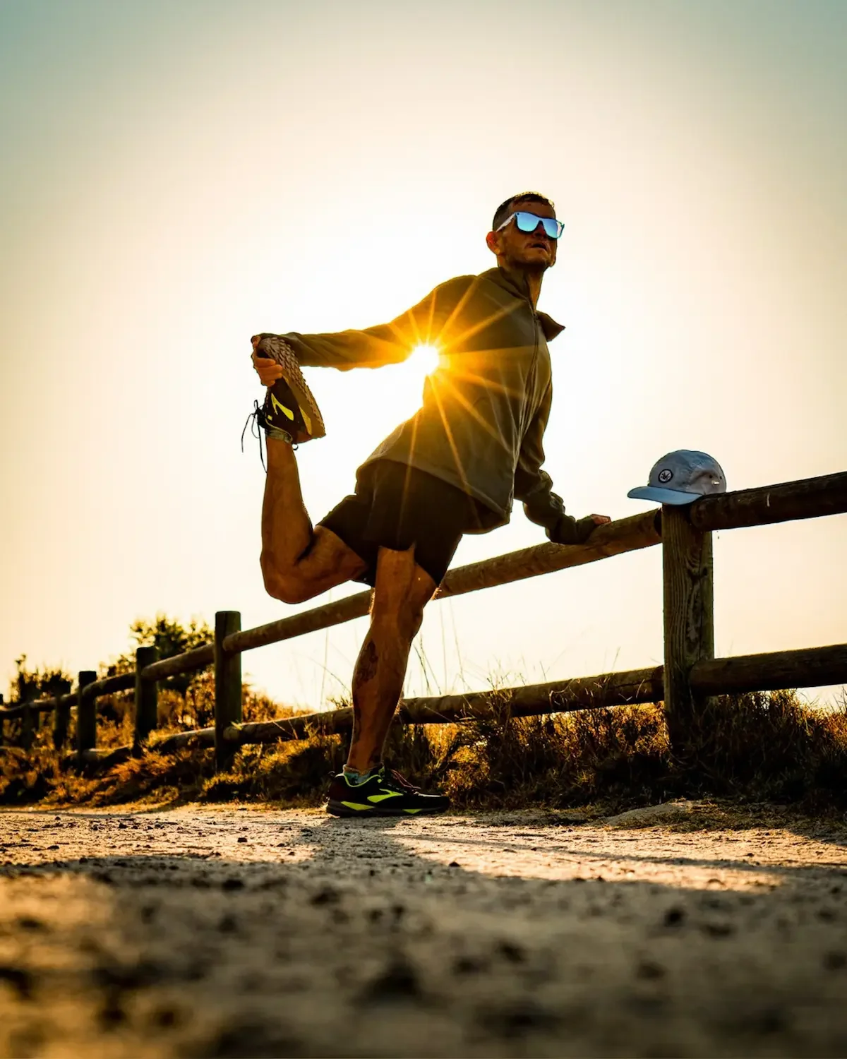 A man in athletic clothing performing a standing yoga pose on a trail during sunset, wearing sunglasses, with a hat resting on the wooden fence nearby.