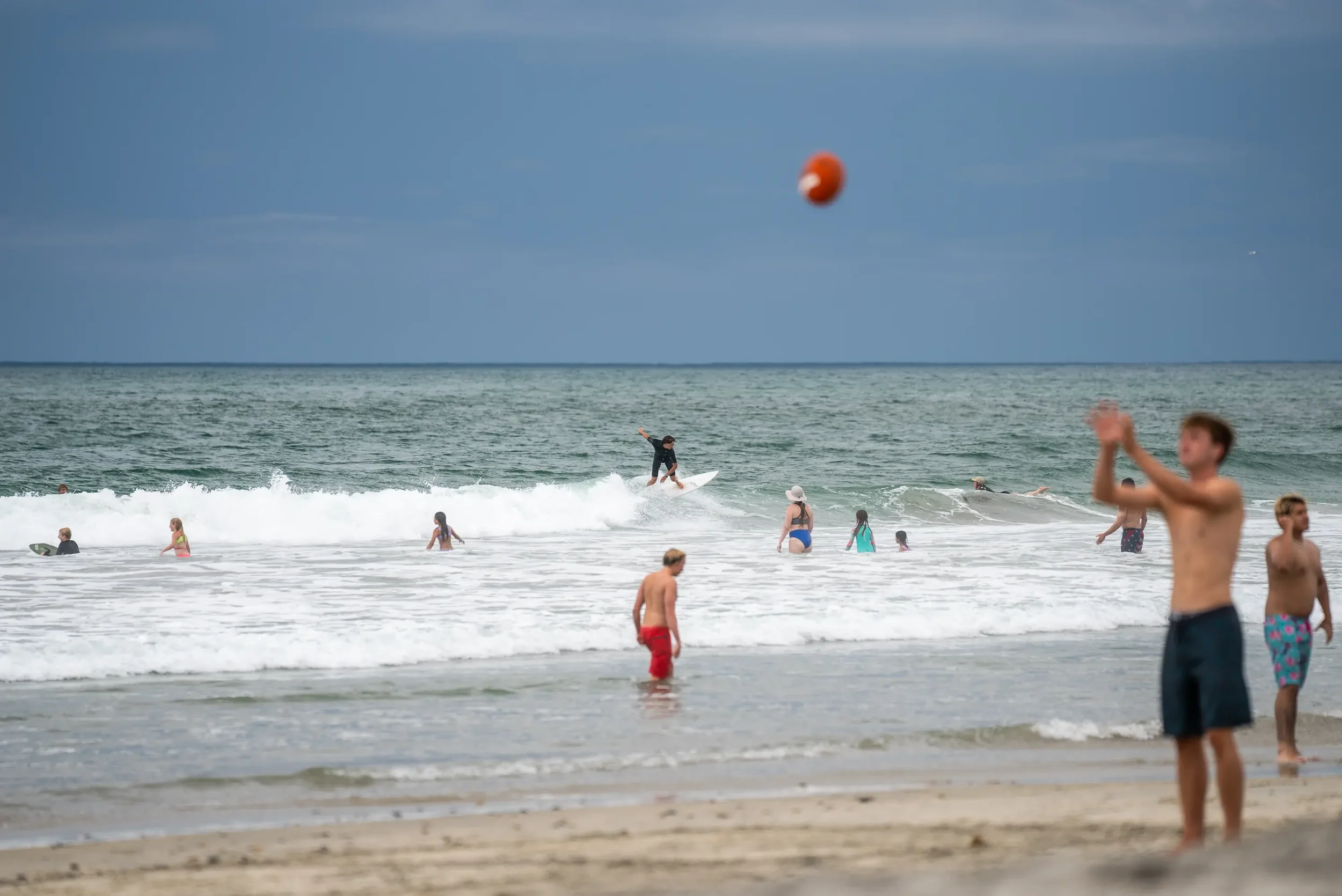 People enjoying a day at the beach with children and adults in the water, some surfing and swimming, under a cloudy sky.