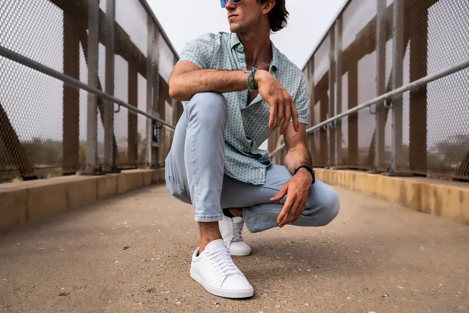 A man crouching on a bridge, wearing white sneakers, light blue jeans, a patterned short-sleeved shirt, and sunglasses, with a cityscape blurred in the background.