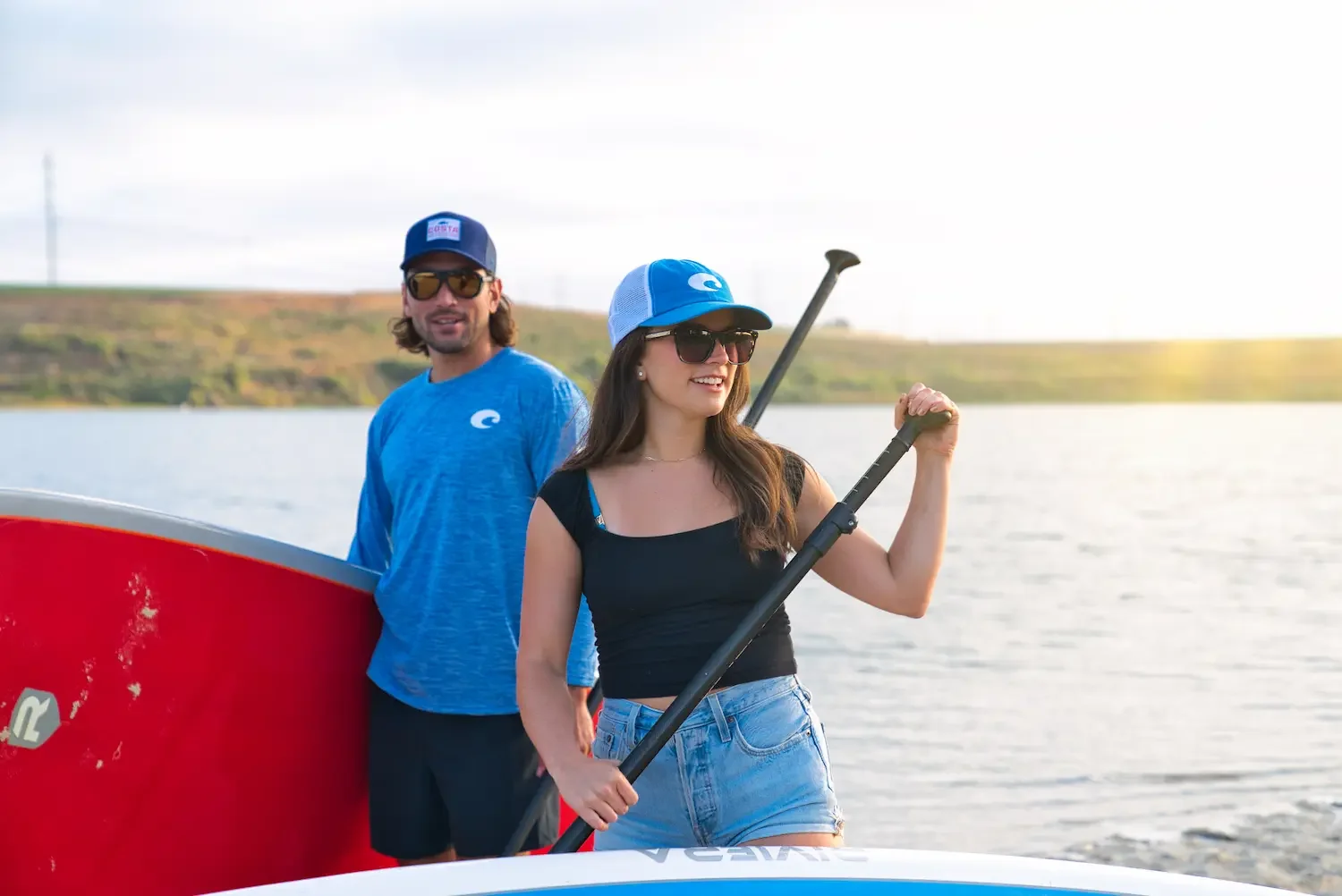 Two young adults, a man and a woman, stand on a boat on a lake with a landscape of hills in the background. The woman holds a paddle, both wearing sunglasses and casual clothing, smiling on a sunny day.