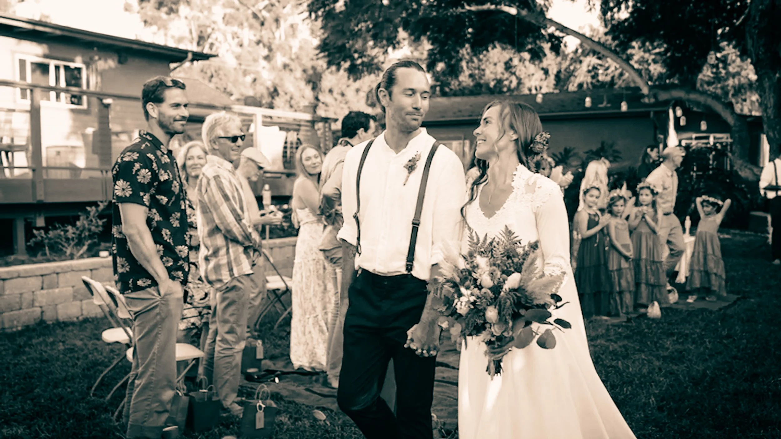 A bride and groom walking together outdoors at a wedding reception, with guests in the background.
