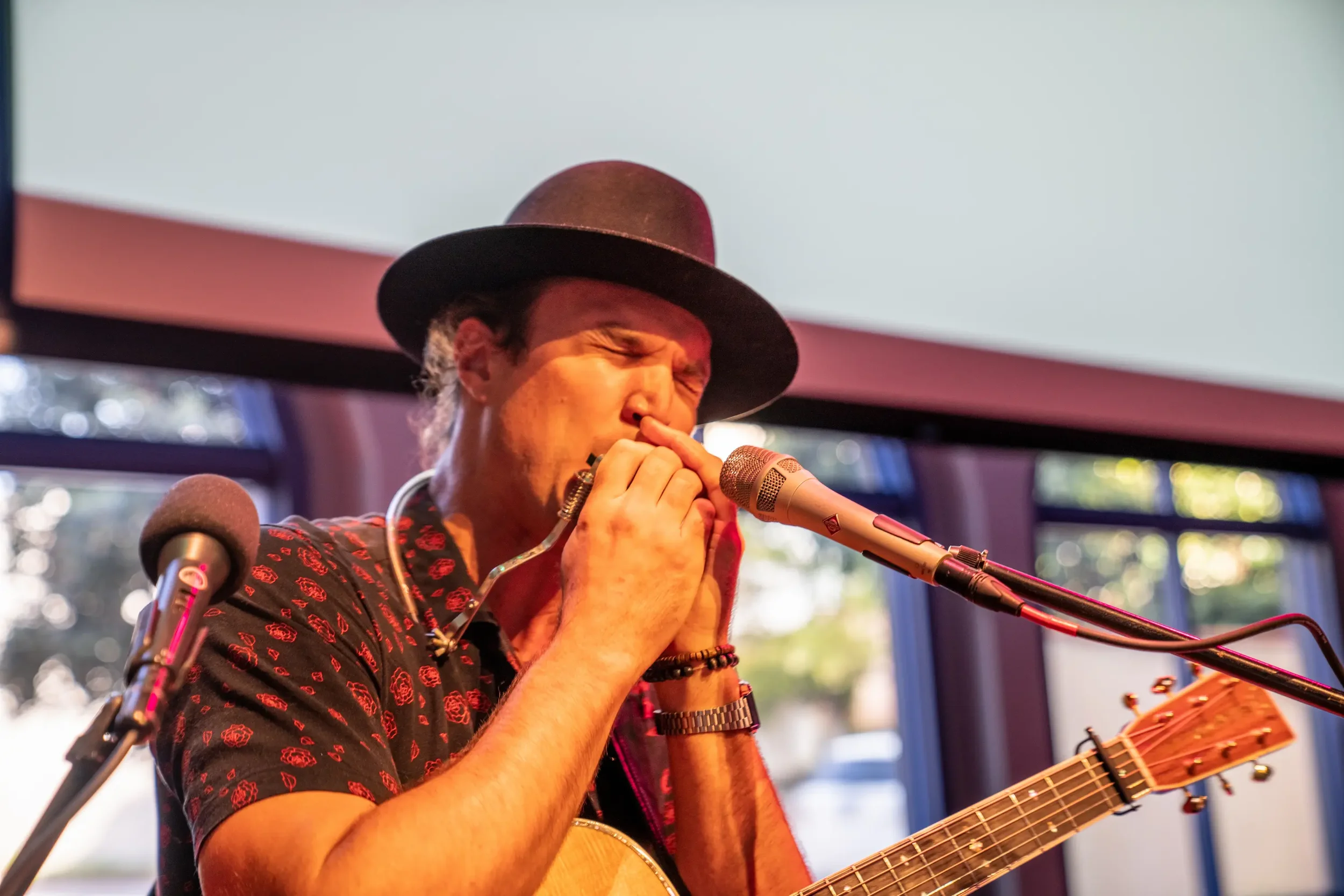A person wearing a black hat and black shirt with red patterns, playing a harmonica while singing into a microphone, with a guitar in hand, during a live performance indoors with a window in the background.