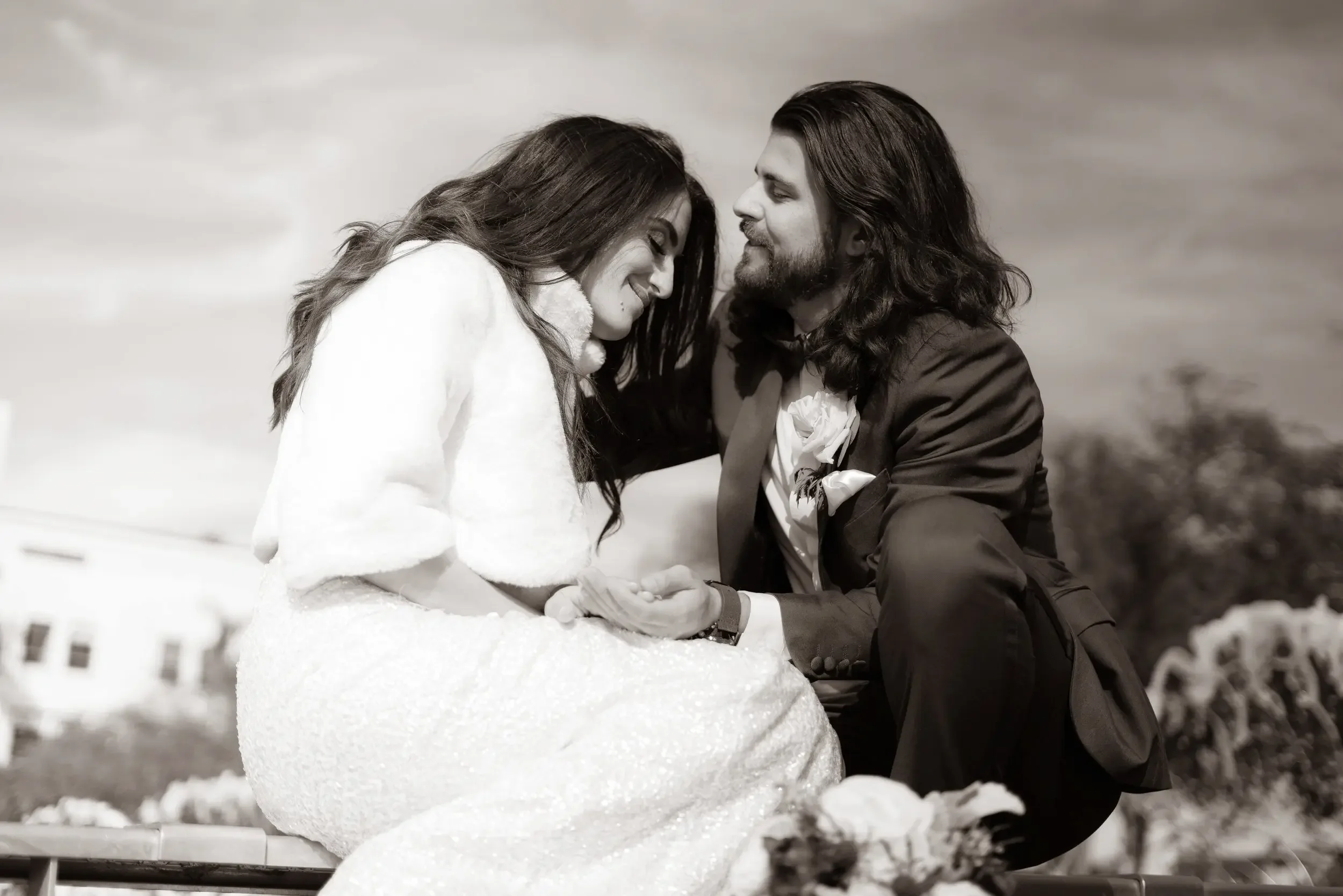 A couple with closed eyes, smiling, sitting close to each other outdoors in winter, holding hands, with snowman in front and a cloudy sky in the background.