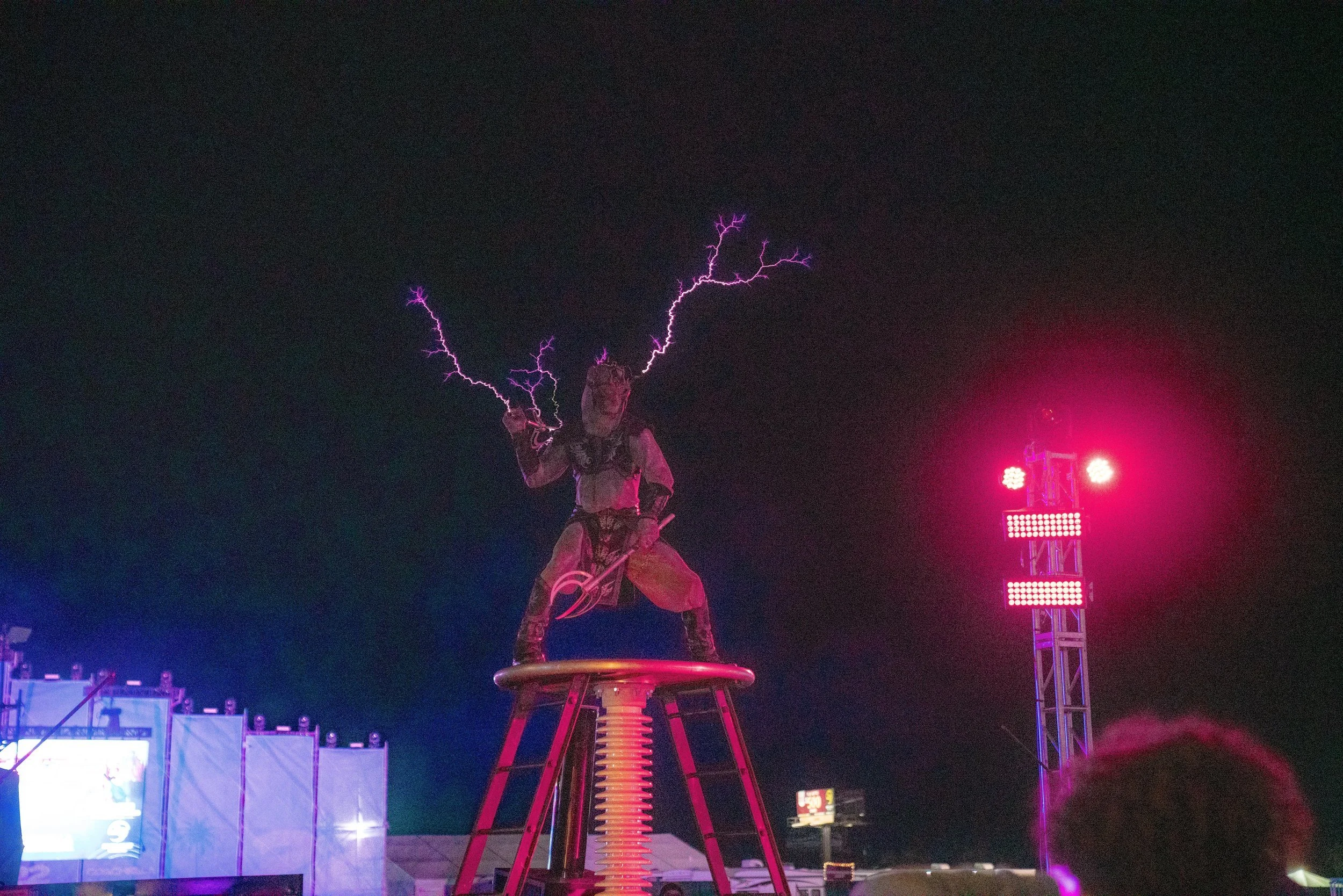 A statue of a man holding what appears to be a tool, with lightning effects above his head, standing on a circular platform at night during a light show.