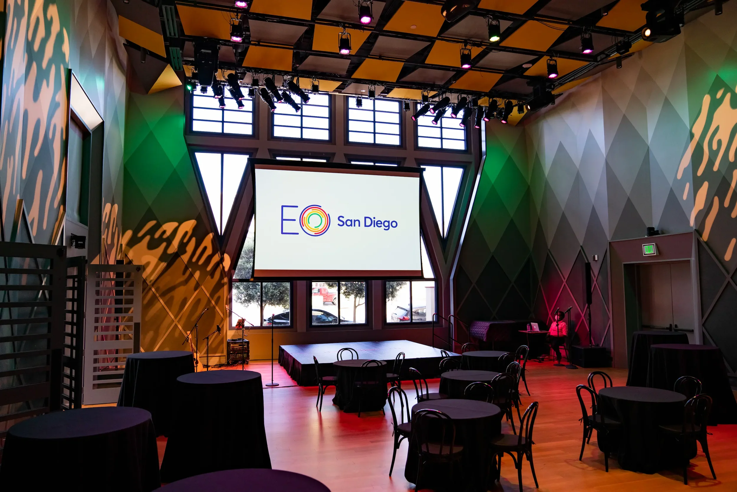 Interior of a conference or event hall with tables and chairs, a large screen displaying 'EO San Diego,' and stage lighting.