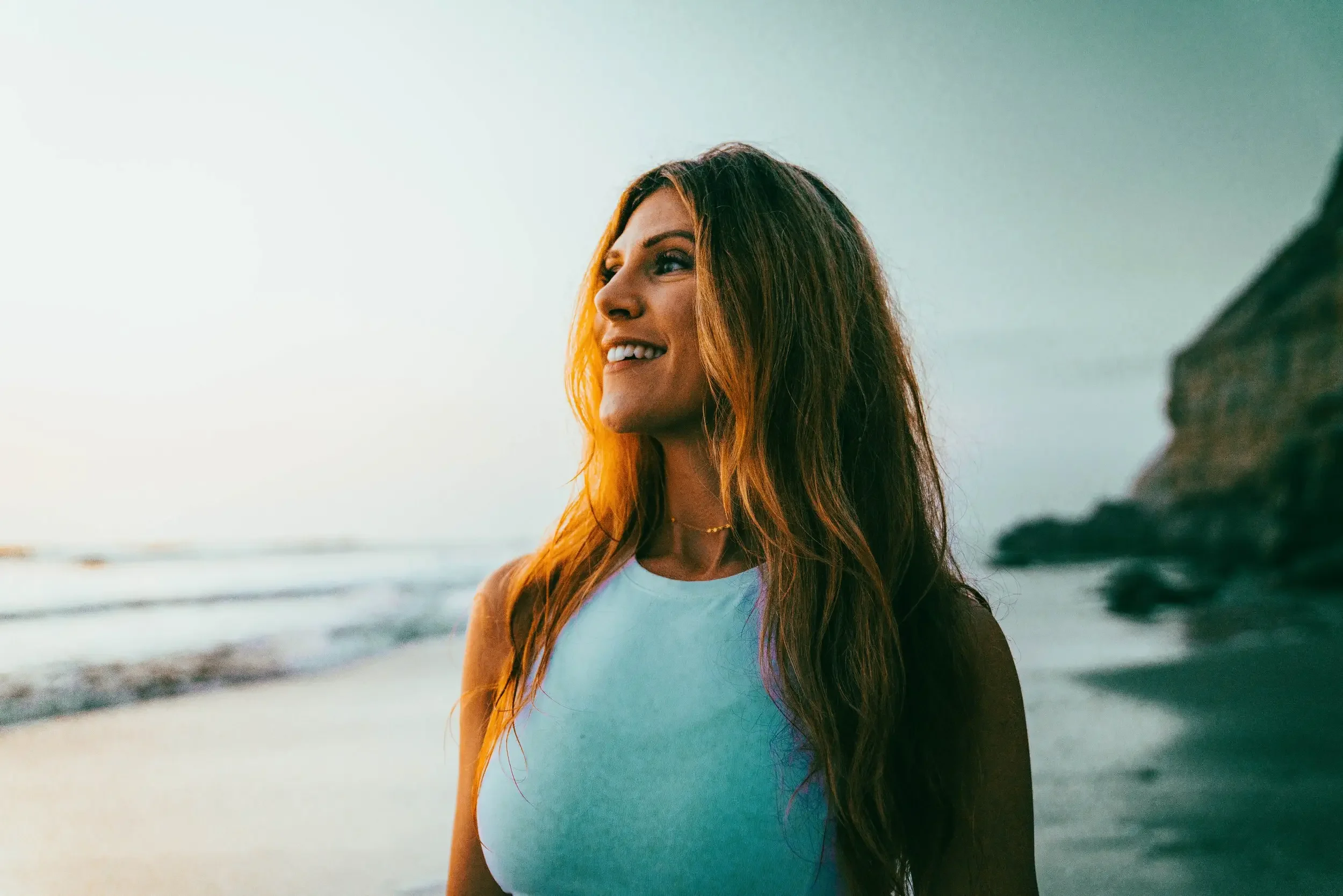 A woman with long, reddish-brown hair smiling while standing on a beach at sunset. She is wearing a white tank top and a delicate necklace, with the ocean and a rocky cliff in the background.