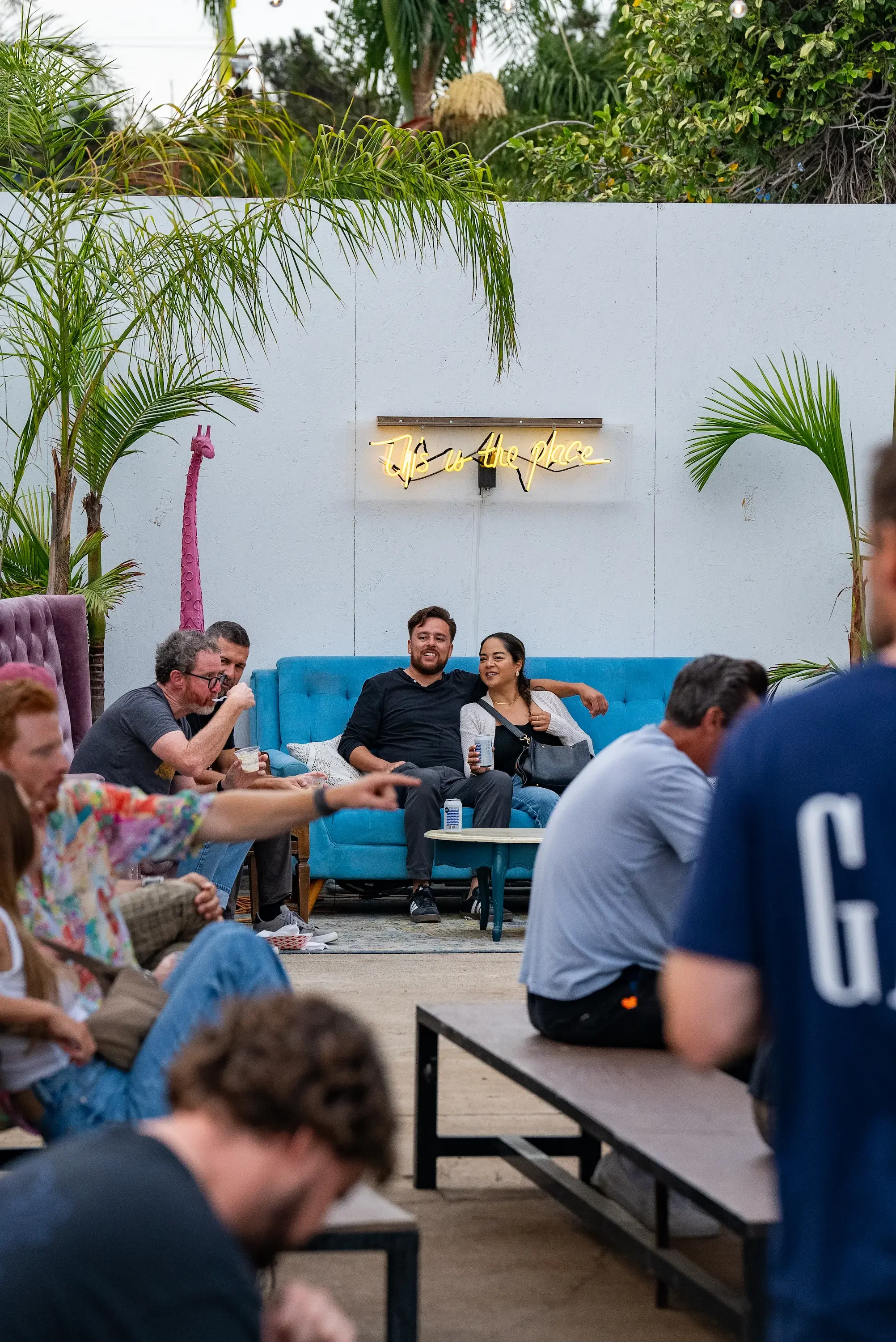 People sitting on the floor and on a couch inside a room with a white wall, green plants, and a neon sign that says 'This is the place'.