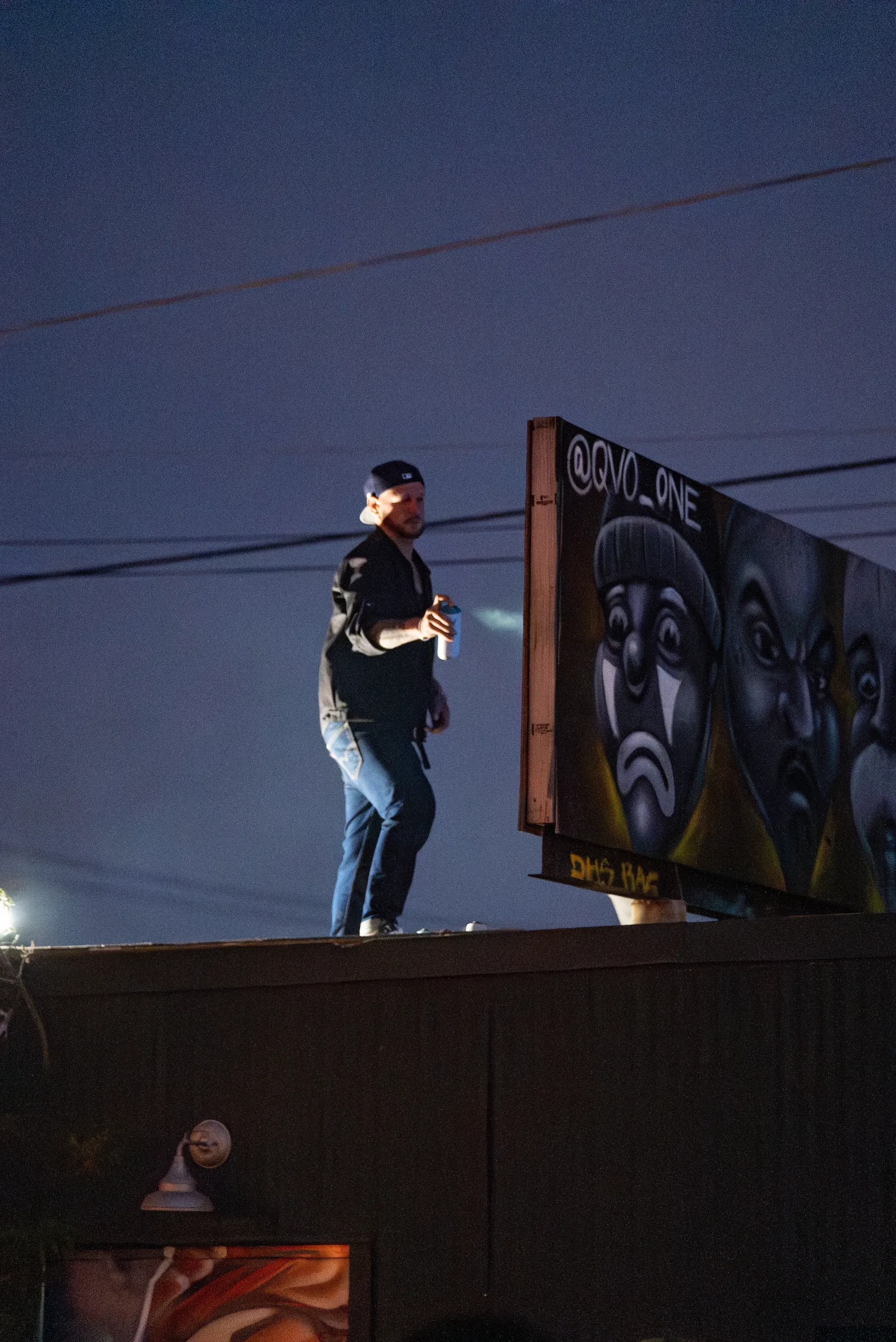 A young man holding a drink stands on top of a building at night, with a billboard featuring African masks and a website handle '@VO_ONE' on it. The sky is dark, and power lines are visible above.