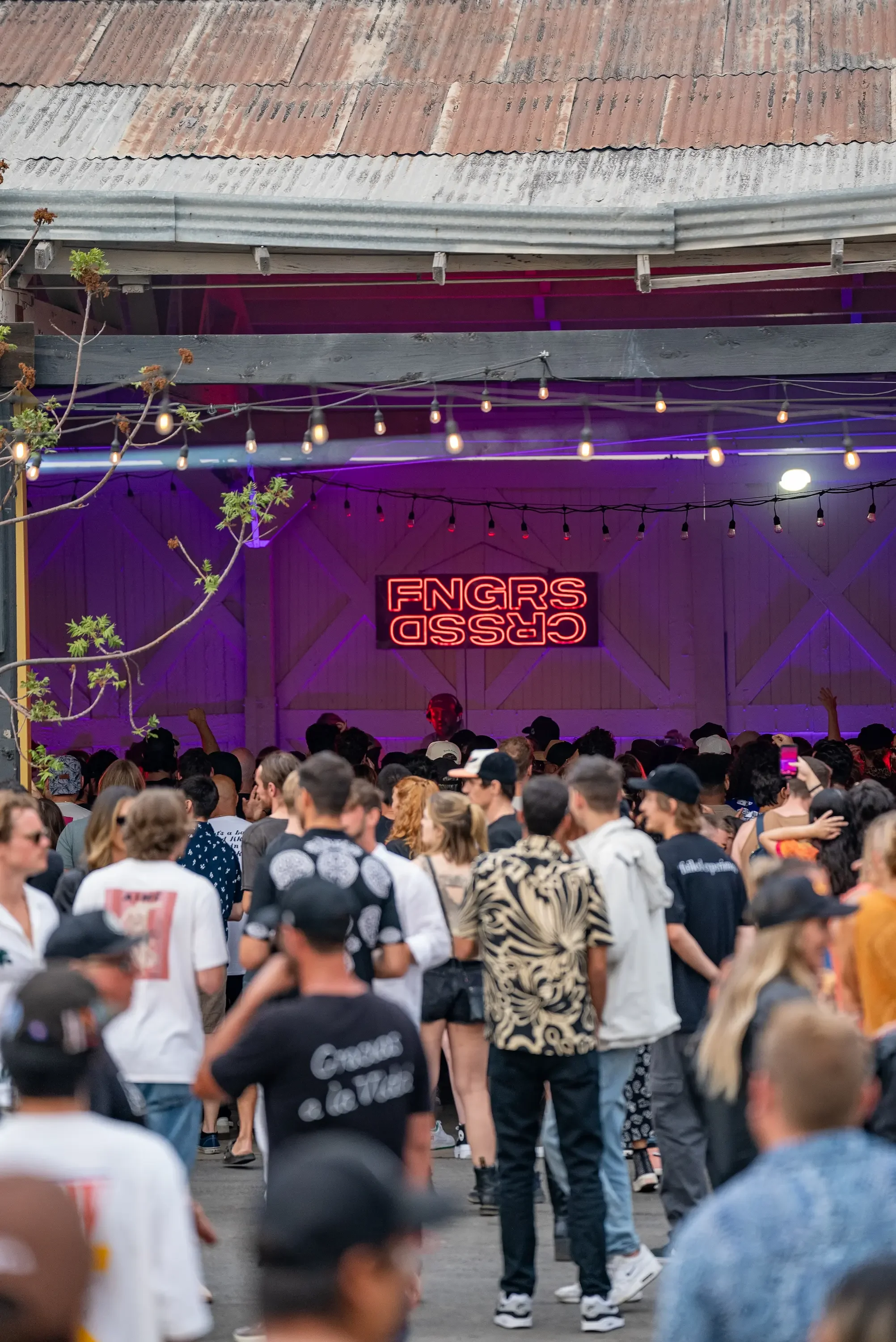 Crowd of people gathered in an outdoor area with a rustic building in the background. The building has a corrugated metal roof, string lights hanging overhead, and a large neon sign that says 'CROSS CHURCH' with the image flipped upside down.