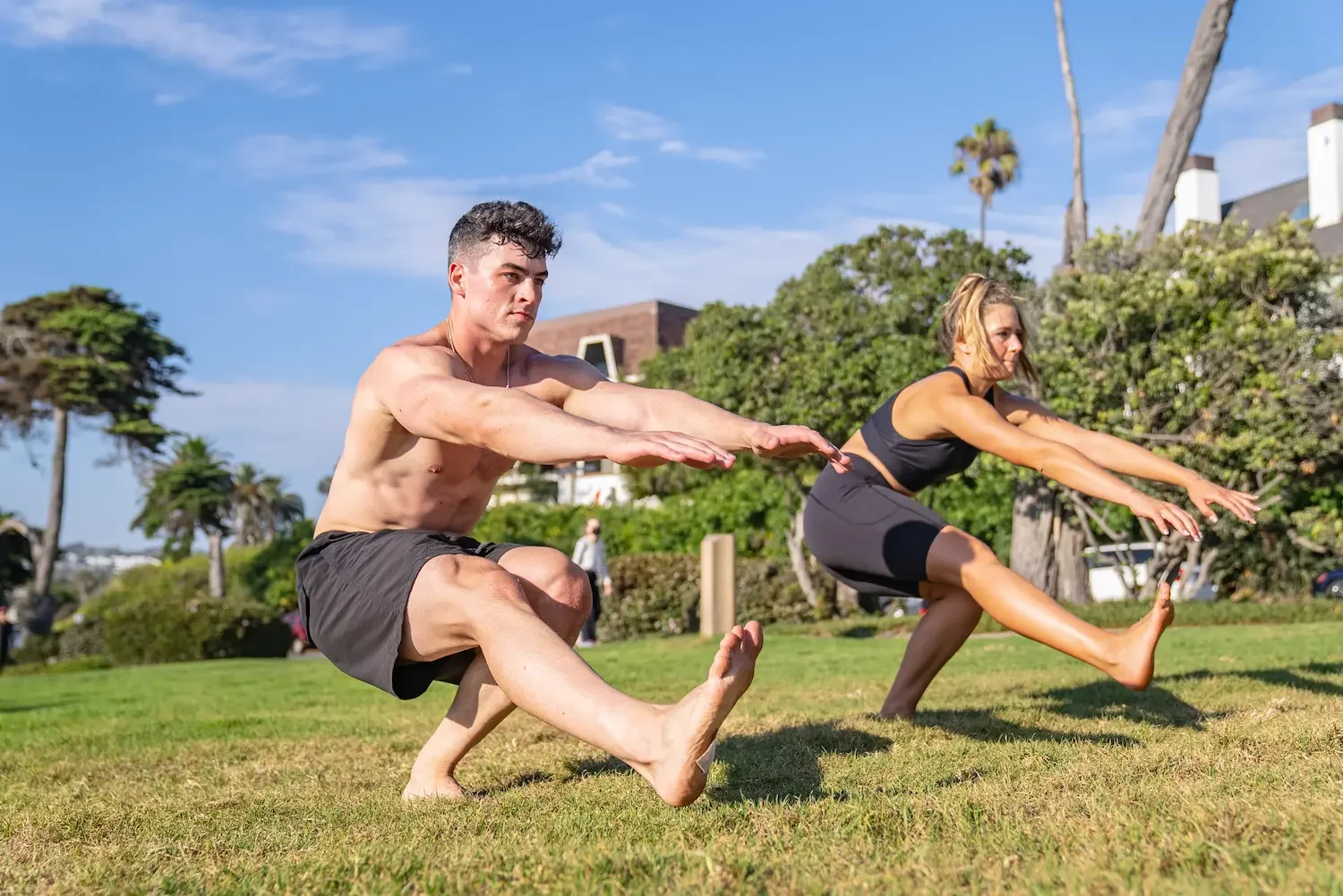 A man and a woman practicing yoga outdoors on a grassy area with trees and buildings in the background. Both are in a balancing pose with extended arms and one leg raised, appearing focused and stable.