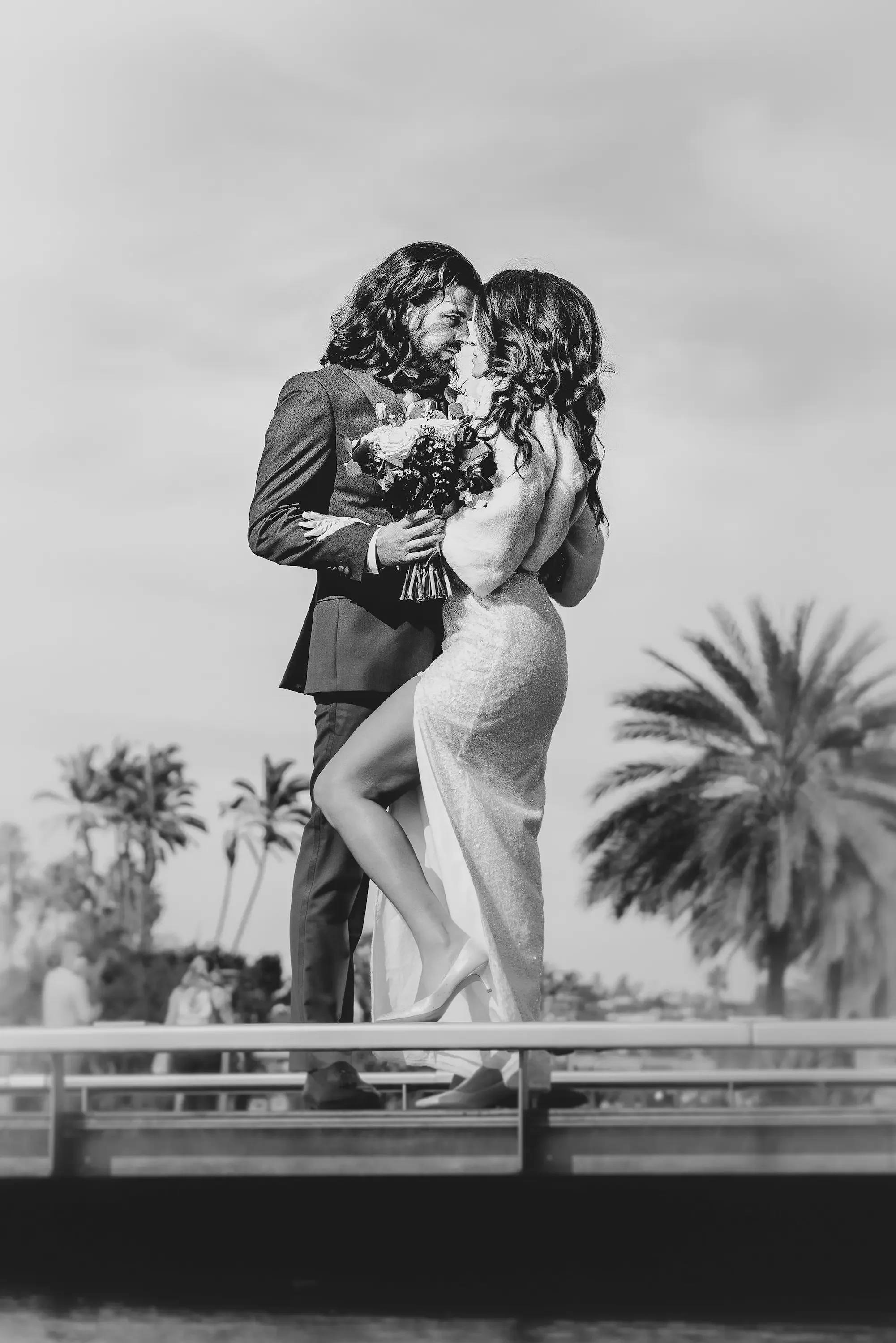 A black and white photo of a couple in wedding attire, embracing each other closely and smiling. The groom is holding a bouquet of flowers. In the background, there are palm trees and a partly cloudy sky.