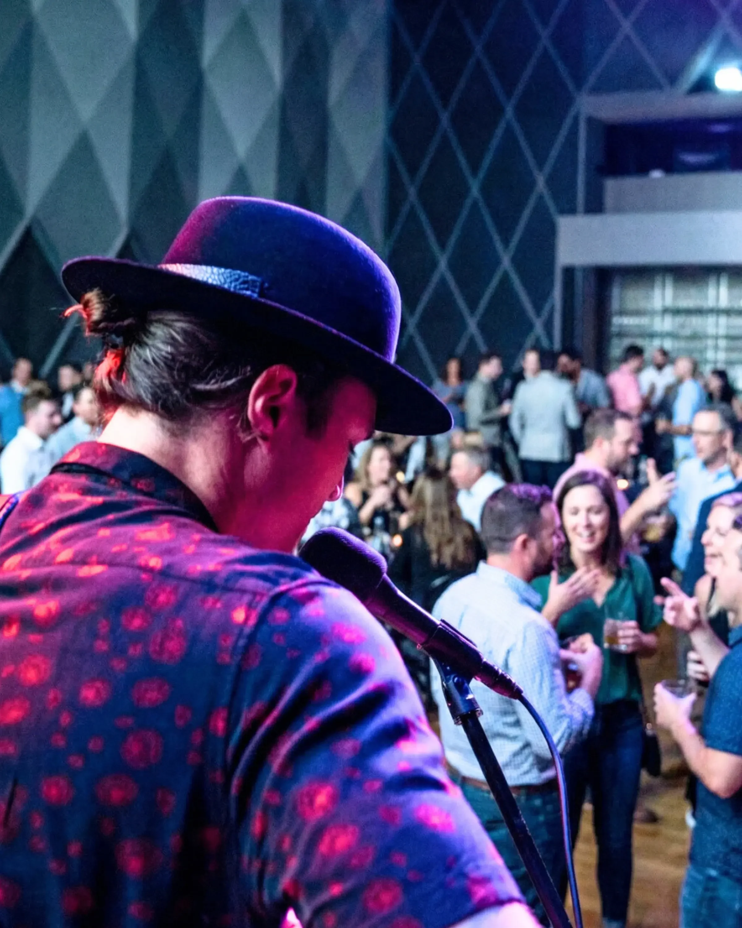 Person wearing a purple hat and a patterned shirt, holding a microphone at a crowded indoor event.