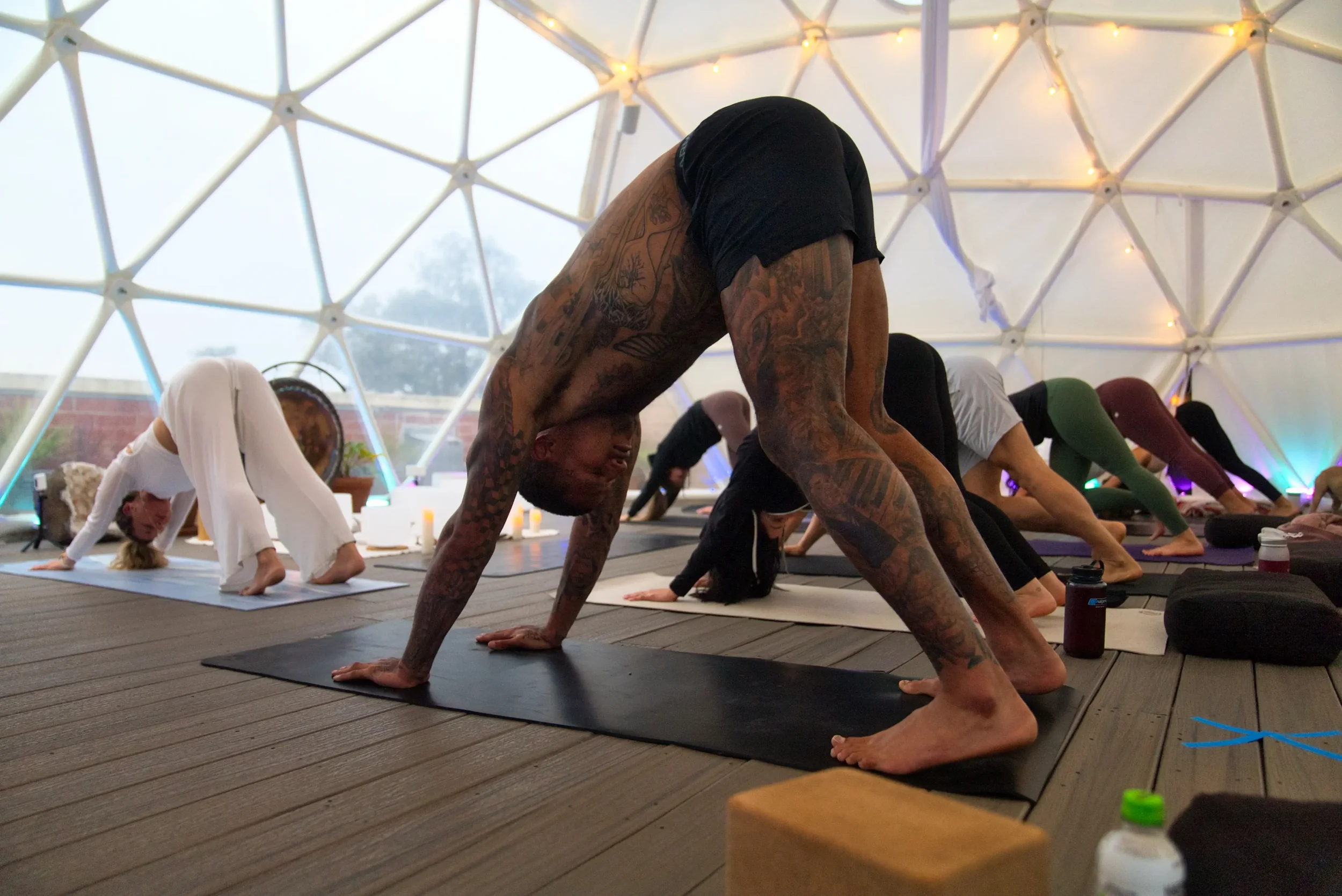 People participating in a yoga class inside a geodesic dome with white walls and ceiling lights. They are in downward dog pose on yoga mats.