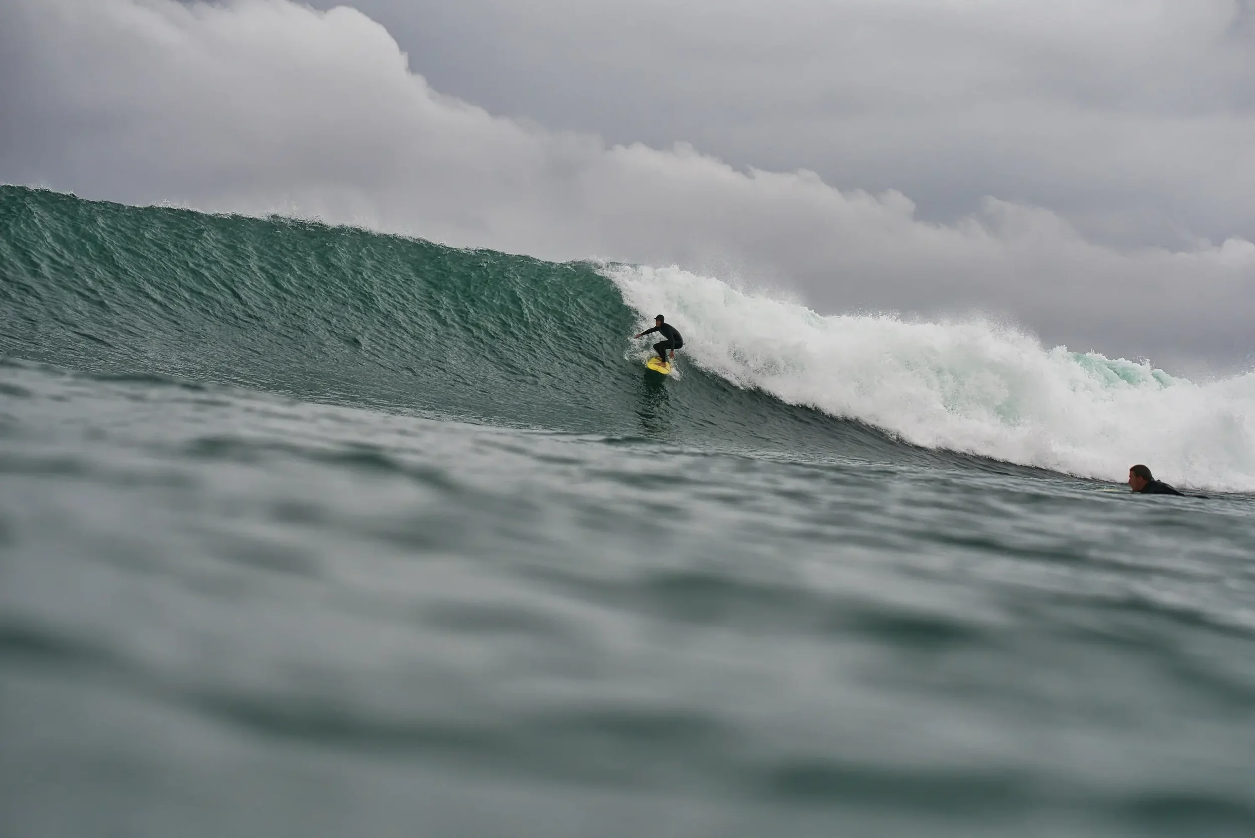A person surfing on a large ocean wave with a cloudy sky in the background.
