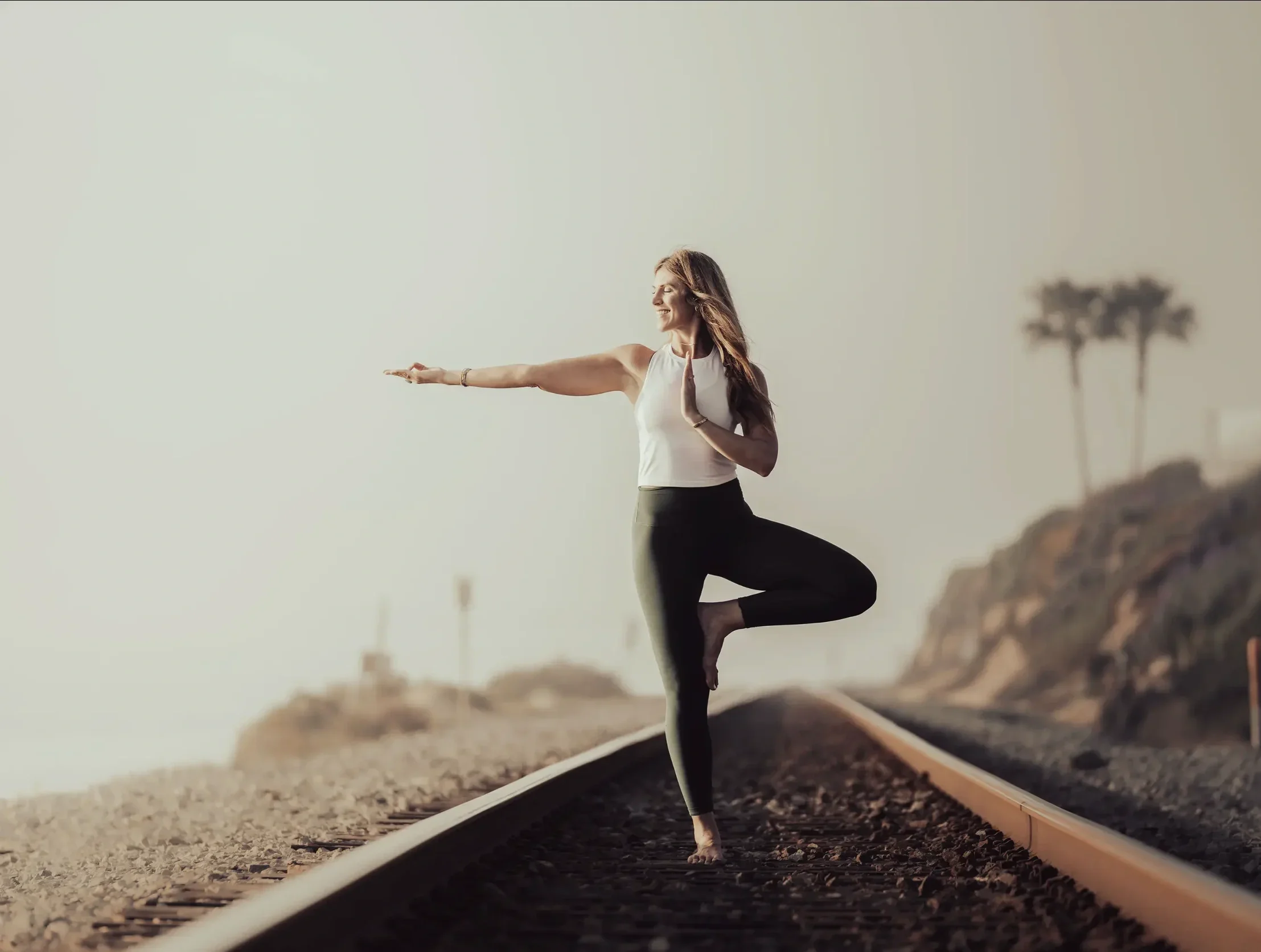 A woman practicing yoga on train tracks outdoors, balancing on one leg with arms extended forward and one hand in a prayer position, during sunset with a blurred background of trees.