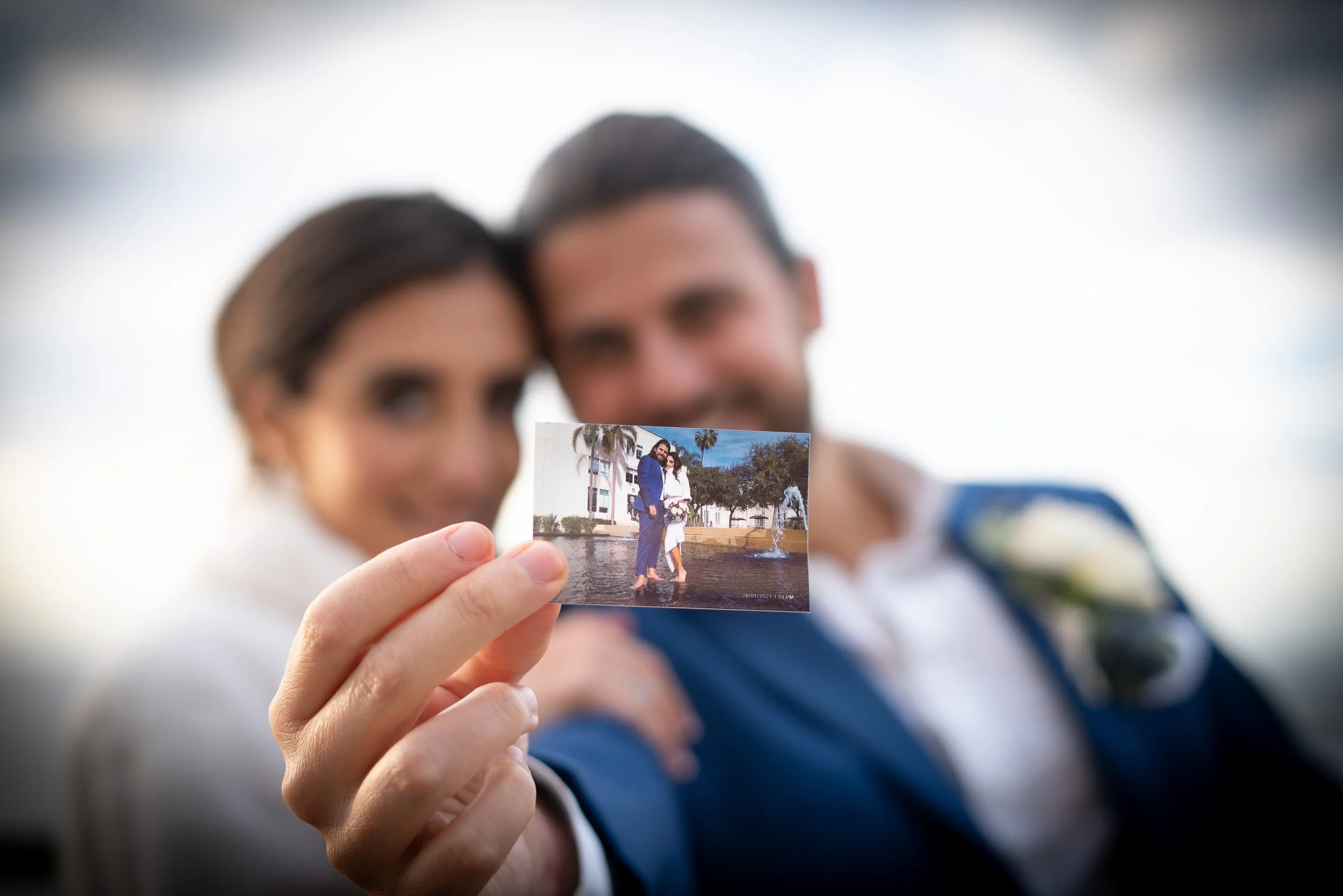 A man and woman hold a small photo in front of their faces, with the background blurred. The photo shows the same couple standing by a fountain, dressed formally, in a city park with palm trees.