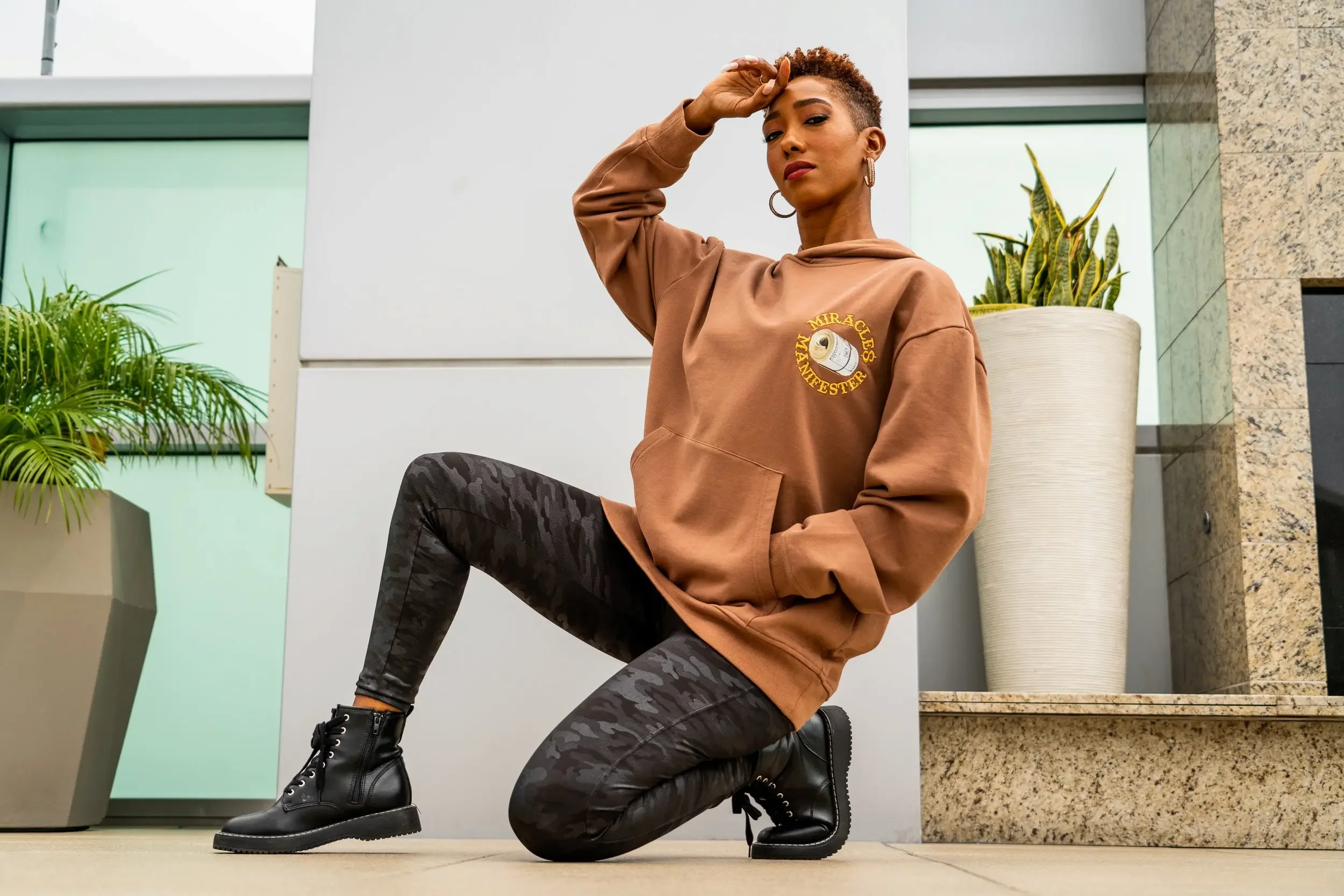 A woman with short curly hair and hoop earrings kneeling indoors, wearing a brown hoodie with a graphic and the words "Miracles Manifest" and black camouflage pants, with one hand on her forehead and the other on her knee, in front of modern white and beige architecture and green plants.