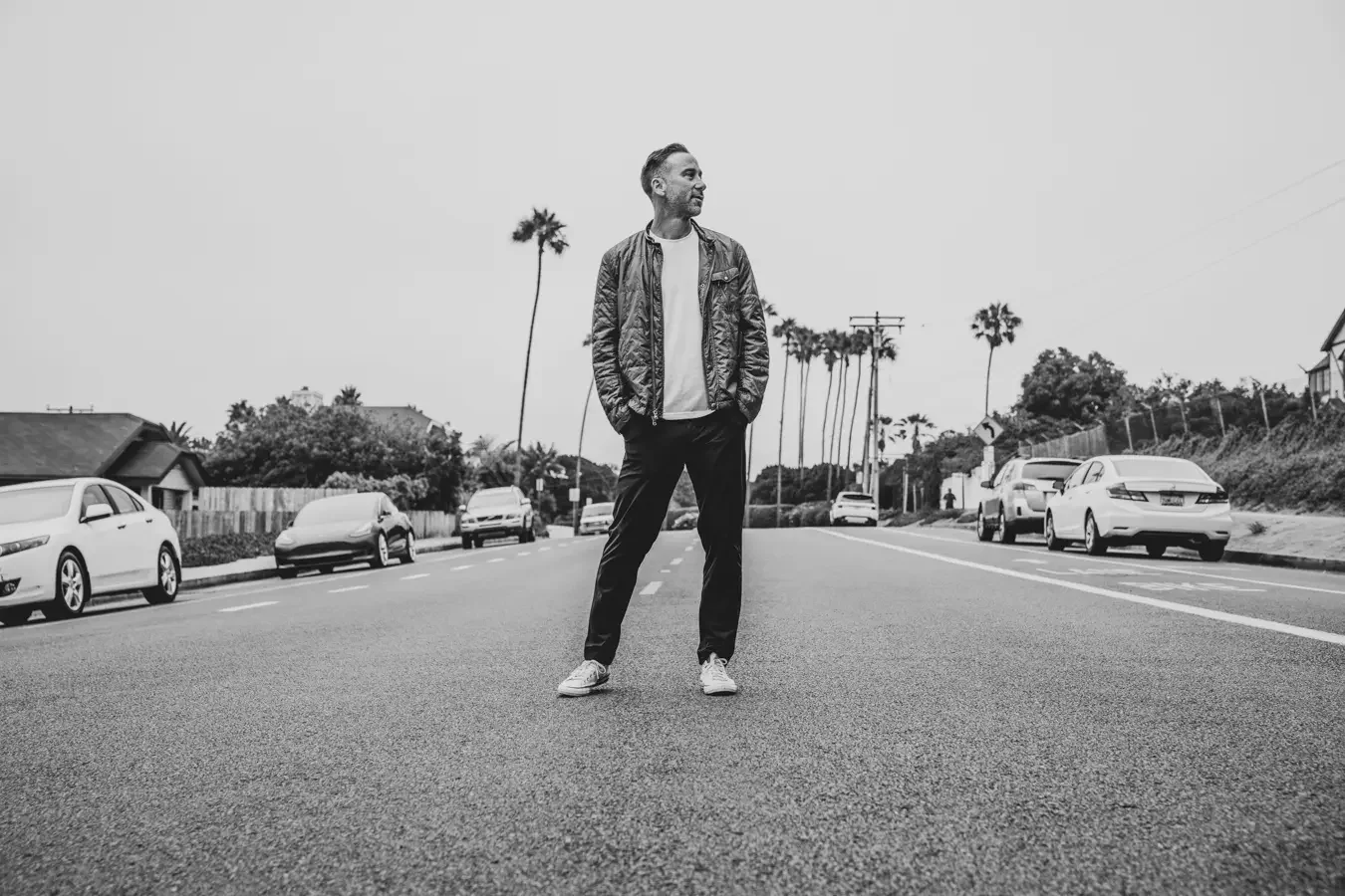 A man stands in the middle of a street with parked cars and palm trees in the background, wearing a jacket, t-shirt, pants, and sneakers.