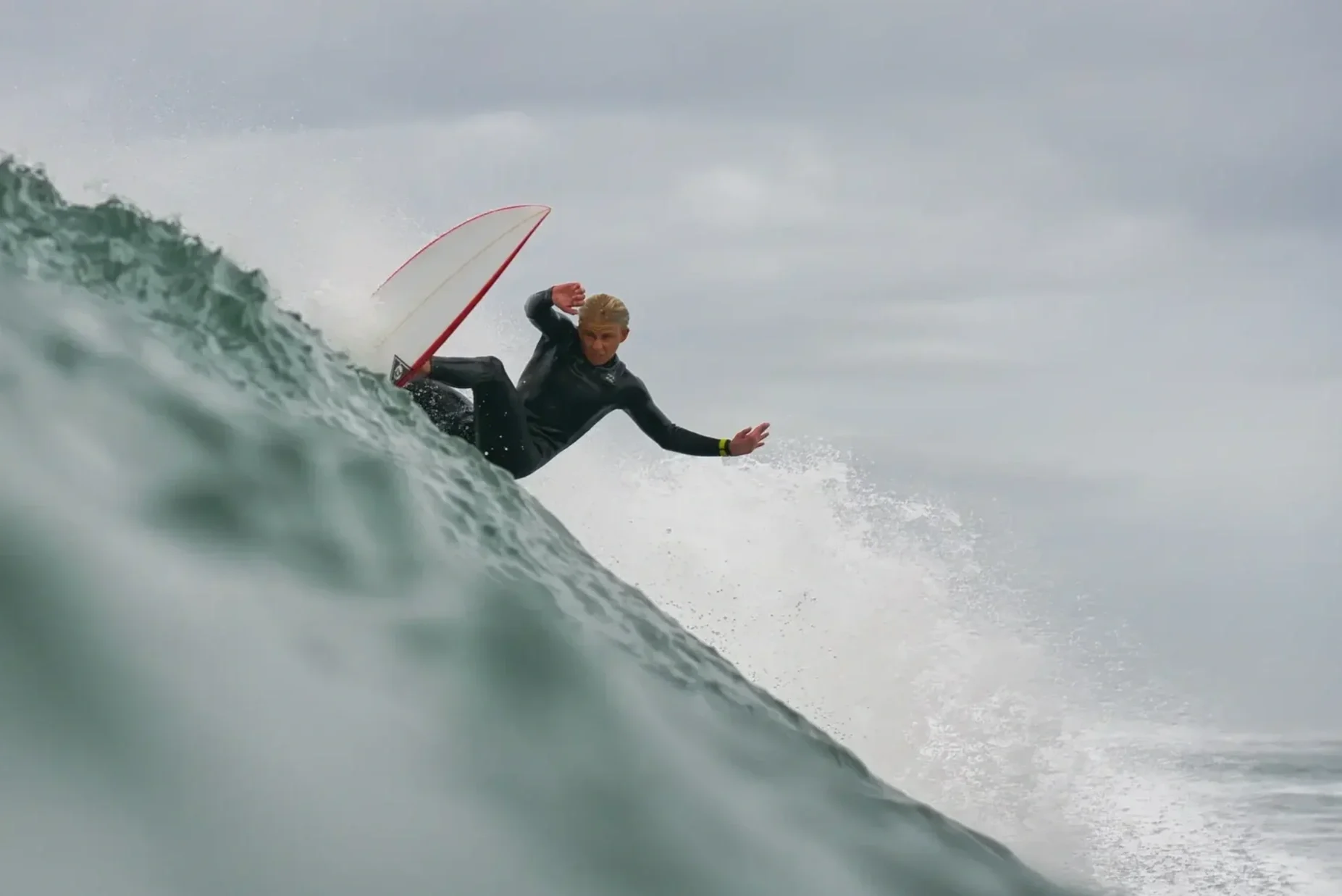 A person with blonde hair surfing on a wave during overcast weather, wearing a black wetsuit and riding a white surfboard with red edges.