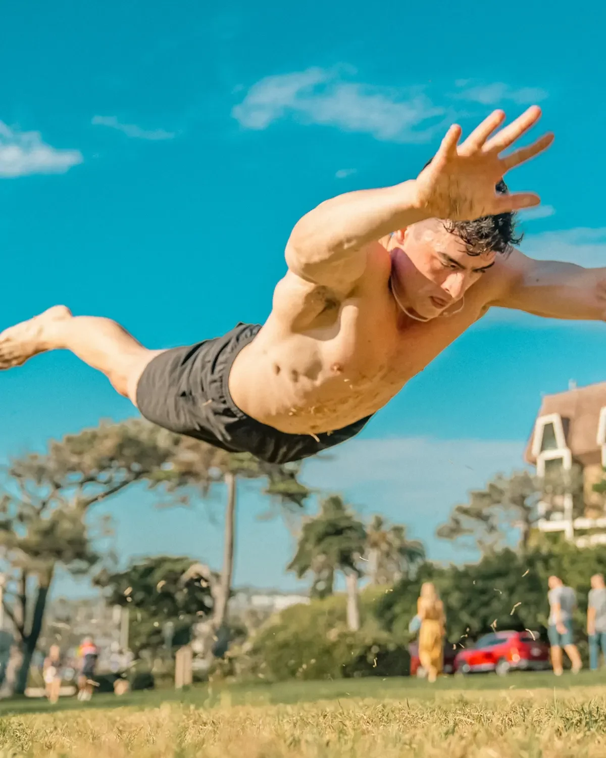 Man jumping into a pool outdoors with trees and buildings in the background under a blue sky.