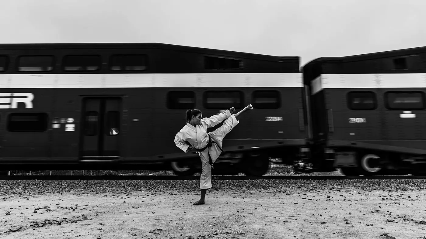 A woman in a karate gi performing a high kick in front of a moving train on a gravel ground