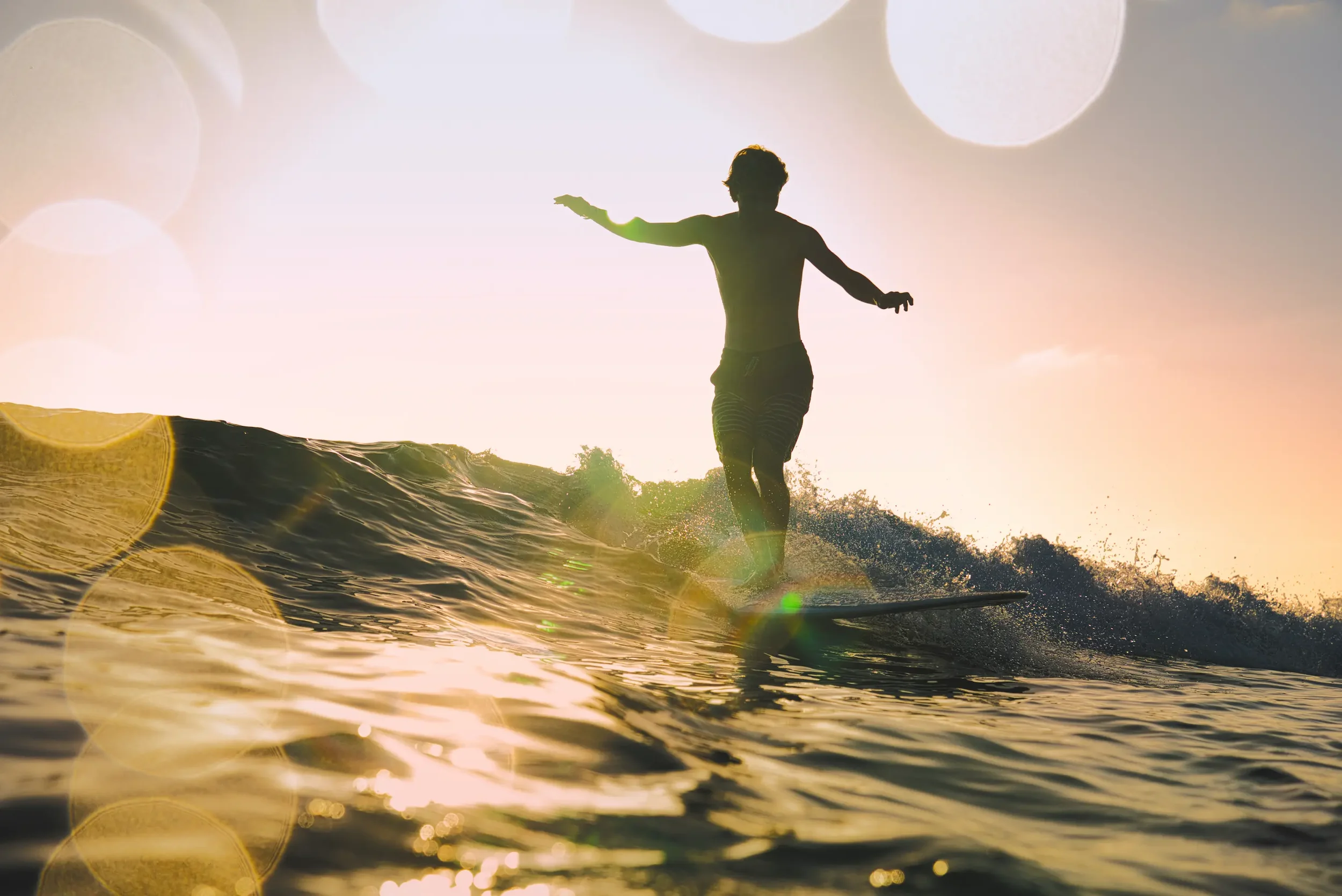 Silhouette of a person surfing on a wave with the sun setting in the background, creating a warm glow and lens flare.